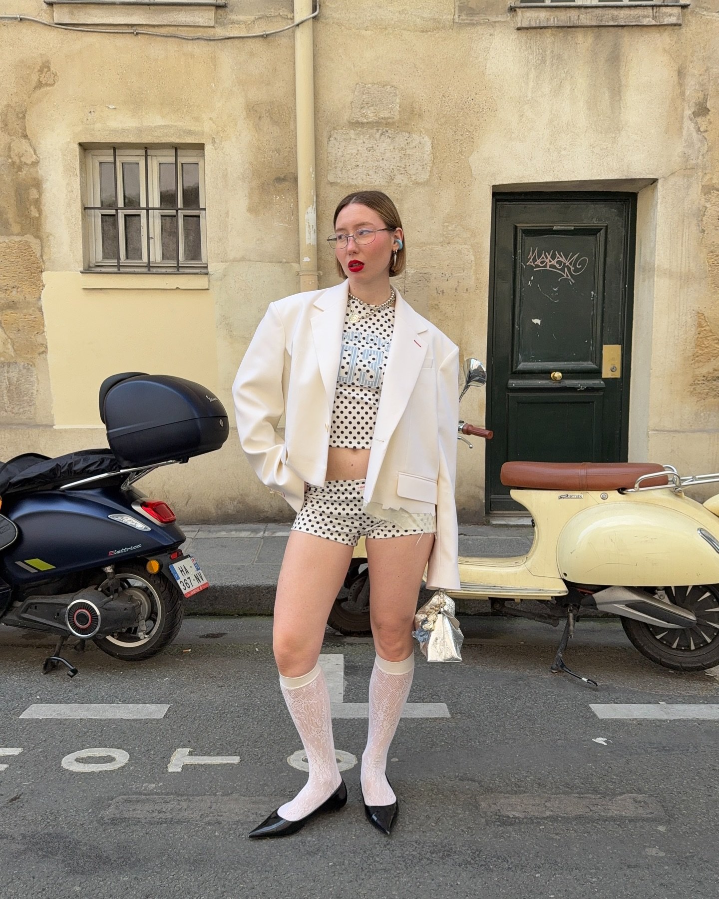 Pessoa em frente a uma parede, vestindo blazer branco e conjunto de top e shorts com estampa de bolinha. Atrás, duas scooters estacionadas.