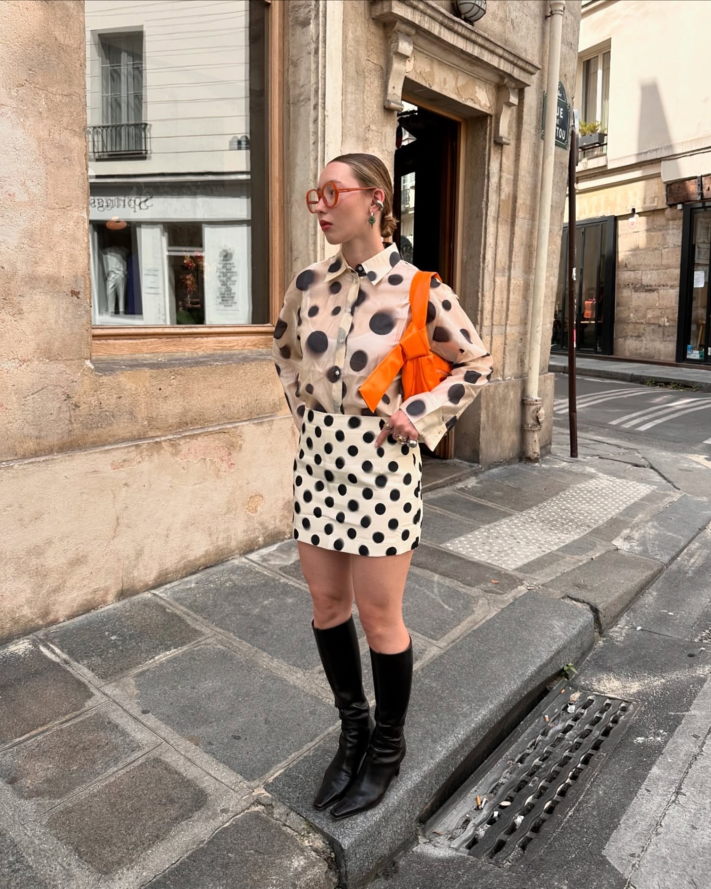 Pessoa em look com estampa de bolinha, usando camisa e saia. Bolsa laranja e botas pretas completam o visual.