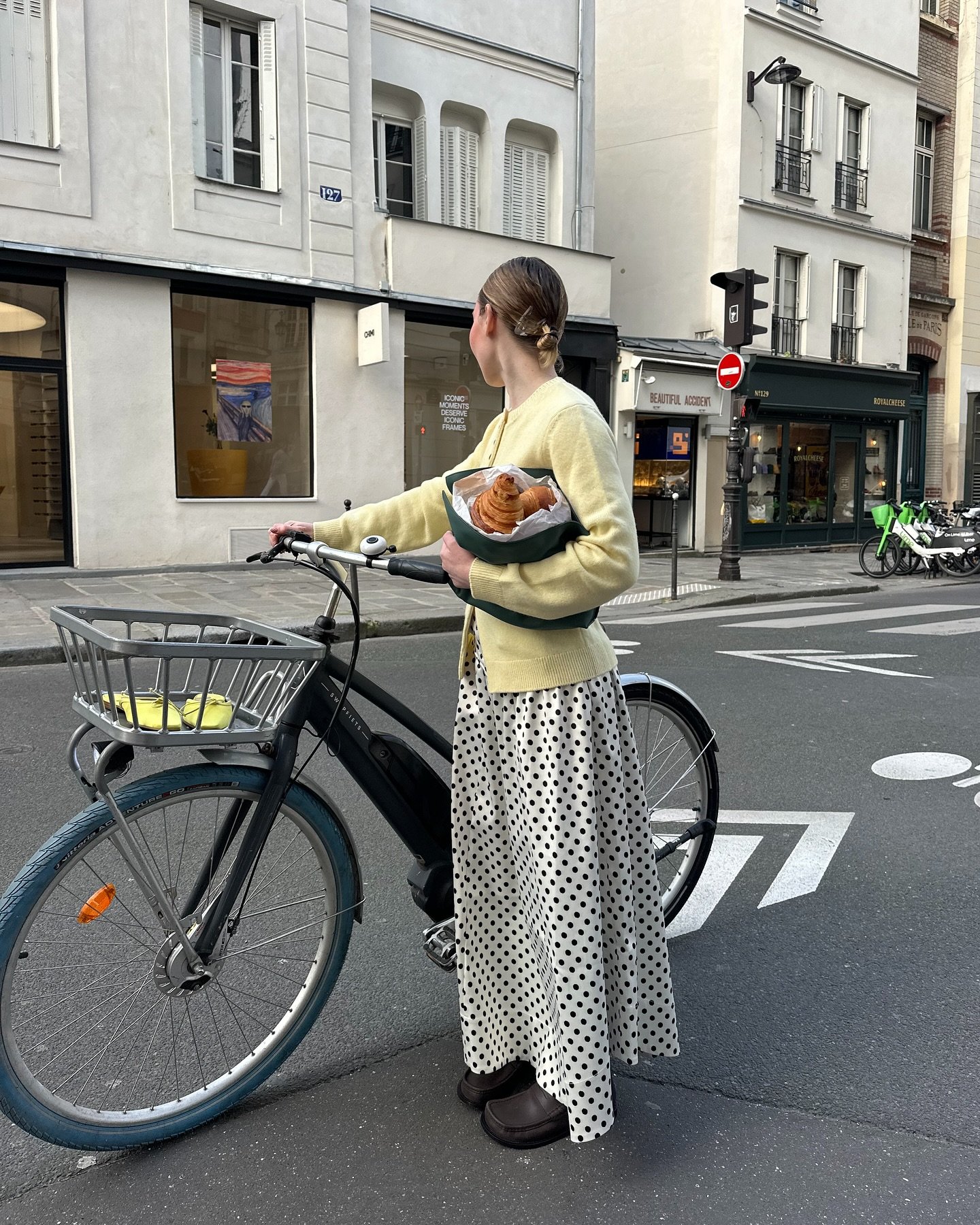Pessoa com saia de estampa de bolinha segura bicicleta e croissants, em rua urbana com prédios atrás.