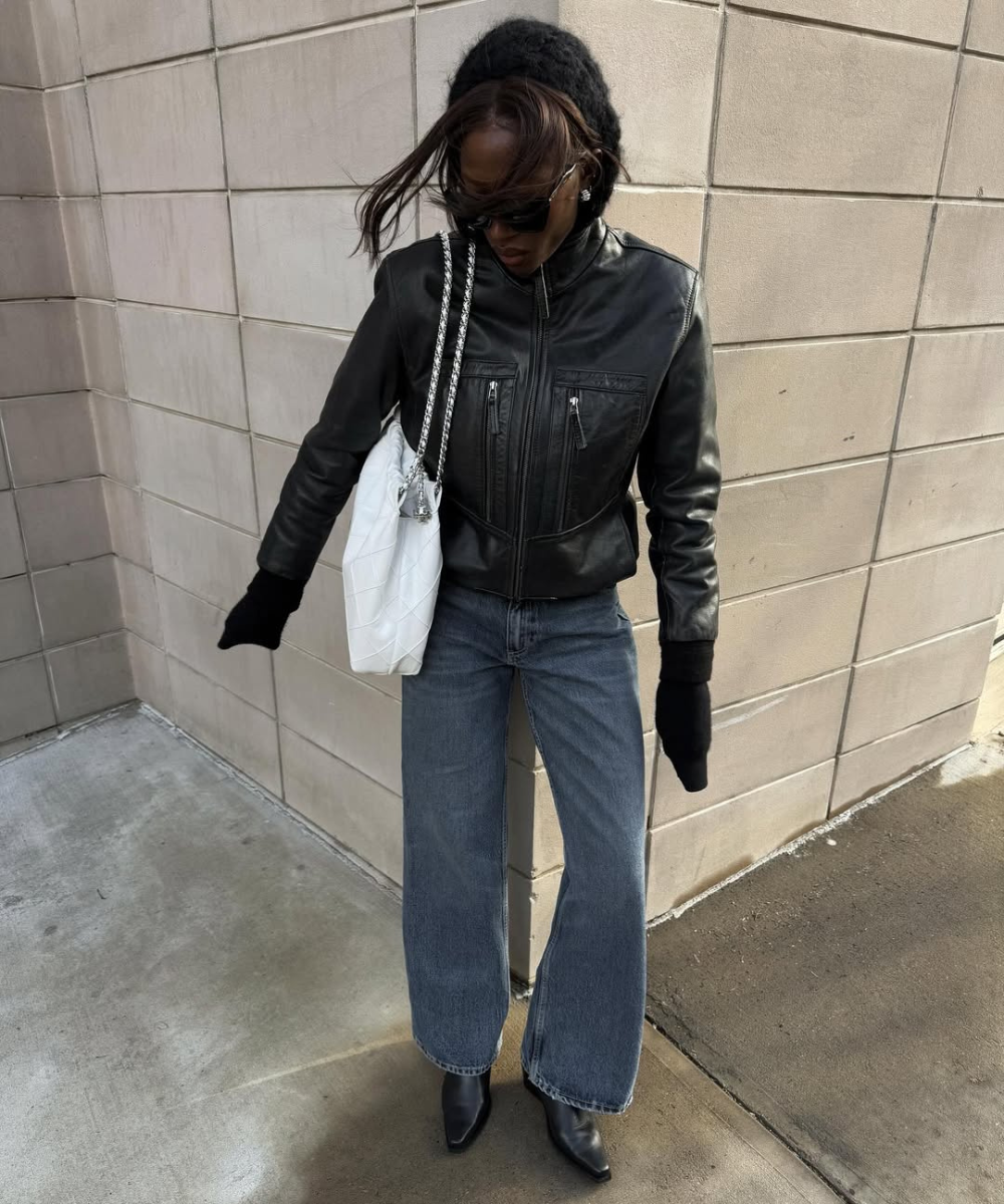 Pessoa com cabelo solto e jaqueta preta, calça jeans, bolsa branca e luvas pretas em cenário urbano.