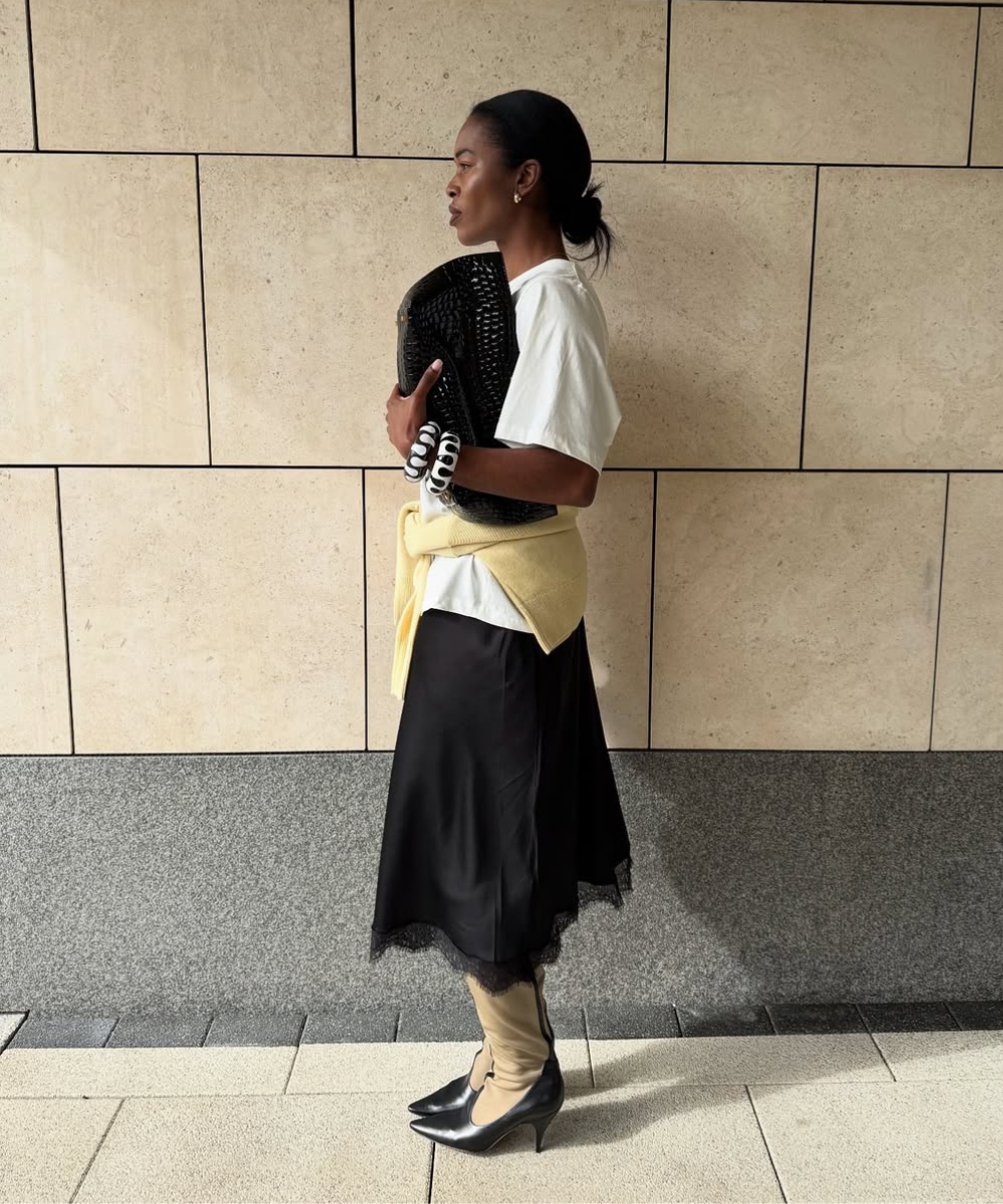 Pessoa com camisa branca e saia preta segurando bolsa preta. Casaco amarelo amarrado na cintura.
