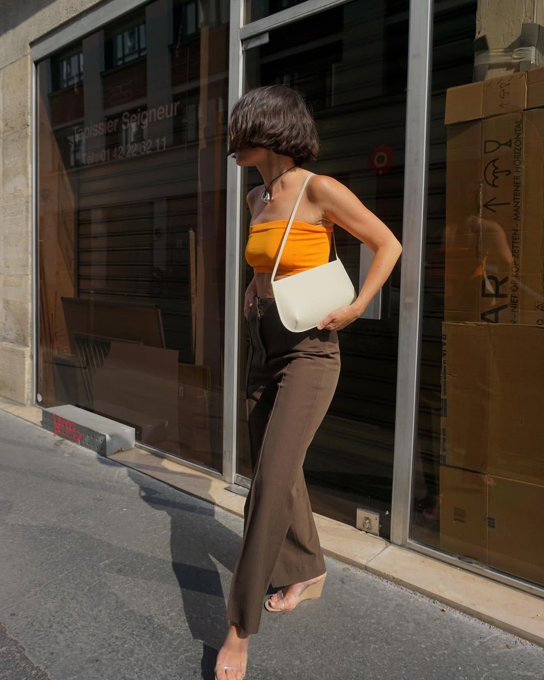 Mulher com top laranja e bolsa branca, usando salto anabela transparente, caminha numa calçada.