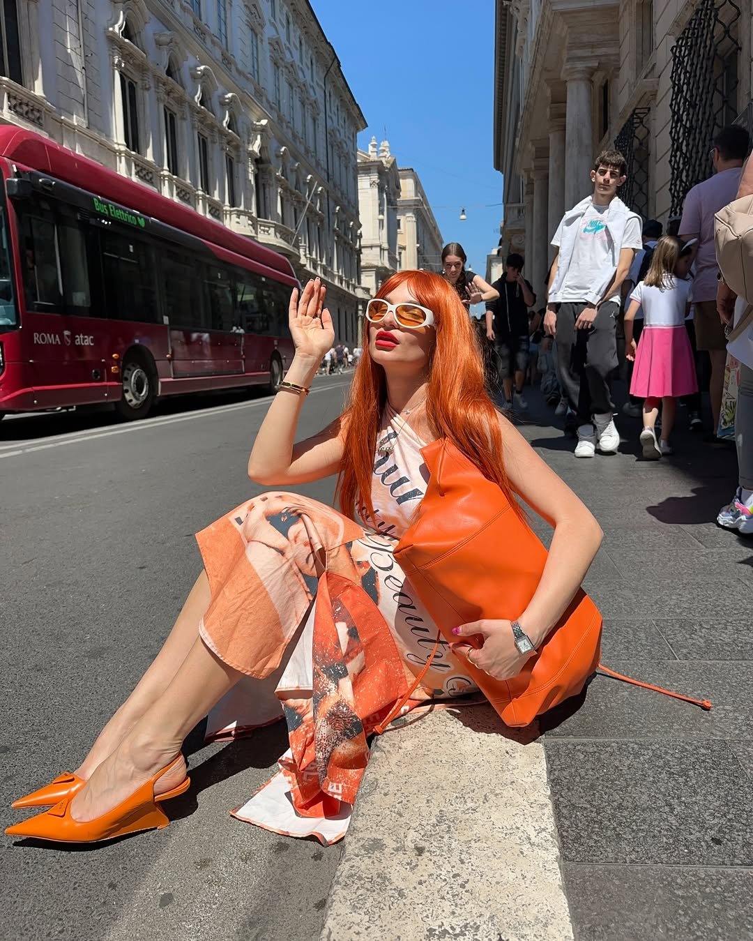 Mulher de cabelo laranja e óculos brancos sentada na calçada com sapatos laranja estilo salto anabela.