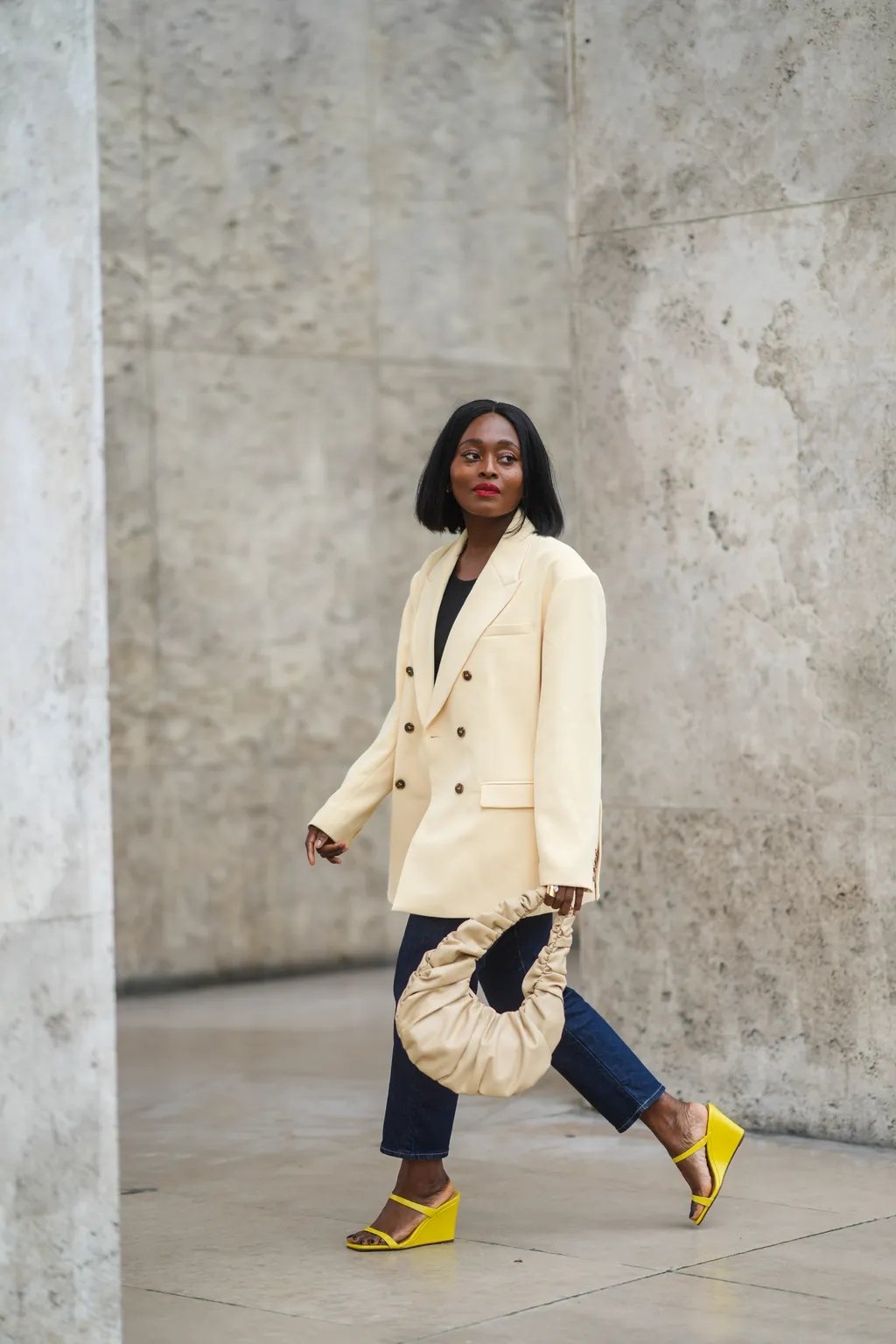 Mulher caminha com blazer claro, calça jeans e sapatos de salto anabela amarelos, segurando bolsa bege.
