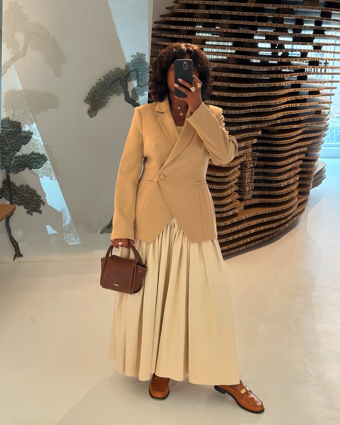Mulher com blazer bege e saia longa, segurando bolsa marrom. Os loafers complementam o look elegante.