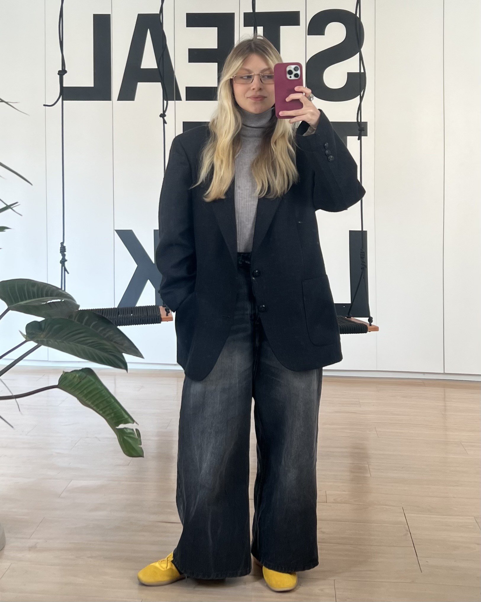 Pessoa de cabelo loiro usa blazer preto e tênis amarelo em frente a um espelho. Plantas ao redor.