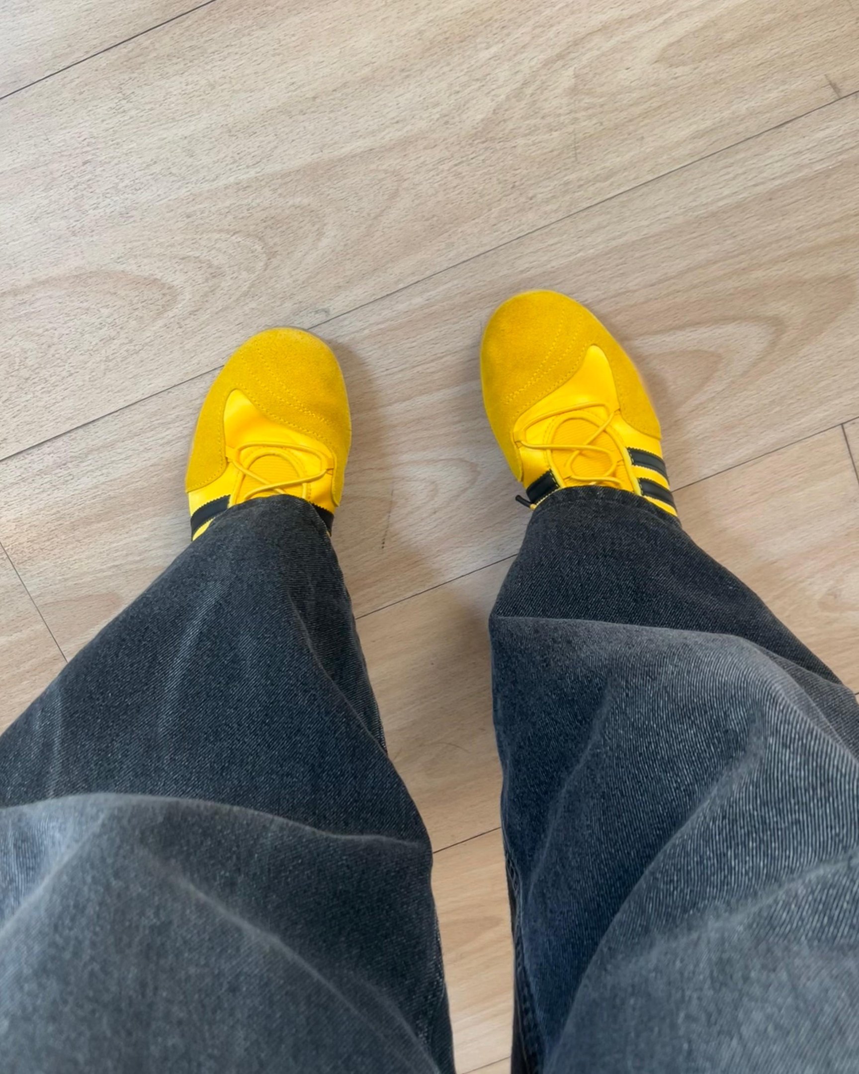 Dois tênis amarelos em um piso de madeira clara, com calça jeans escura.