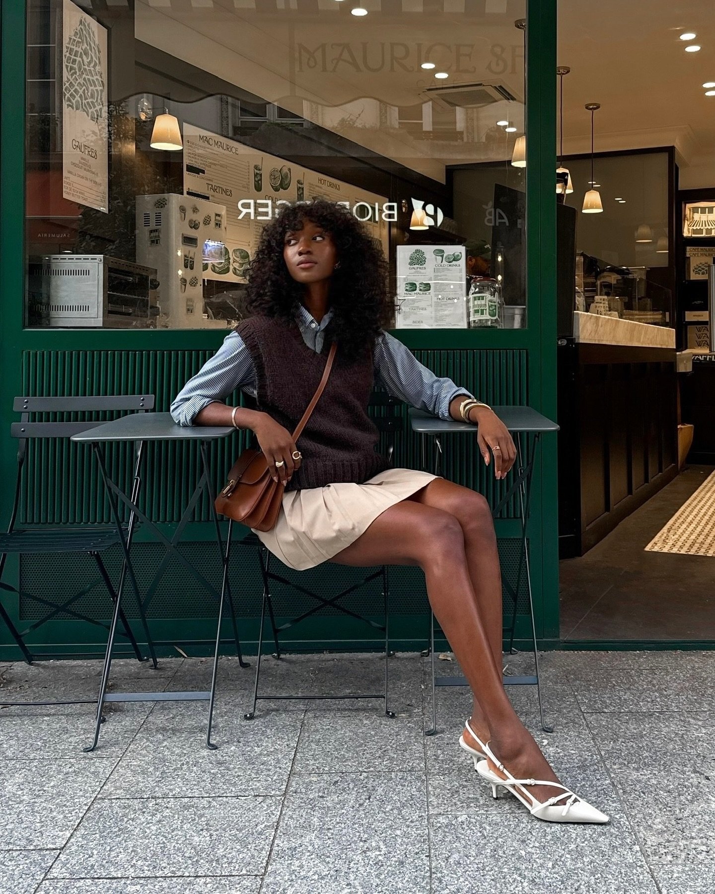 Mulher com colete sobre camisa, minissaia bege e sapatos brancos, sentada em frente a uma cafeteria.