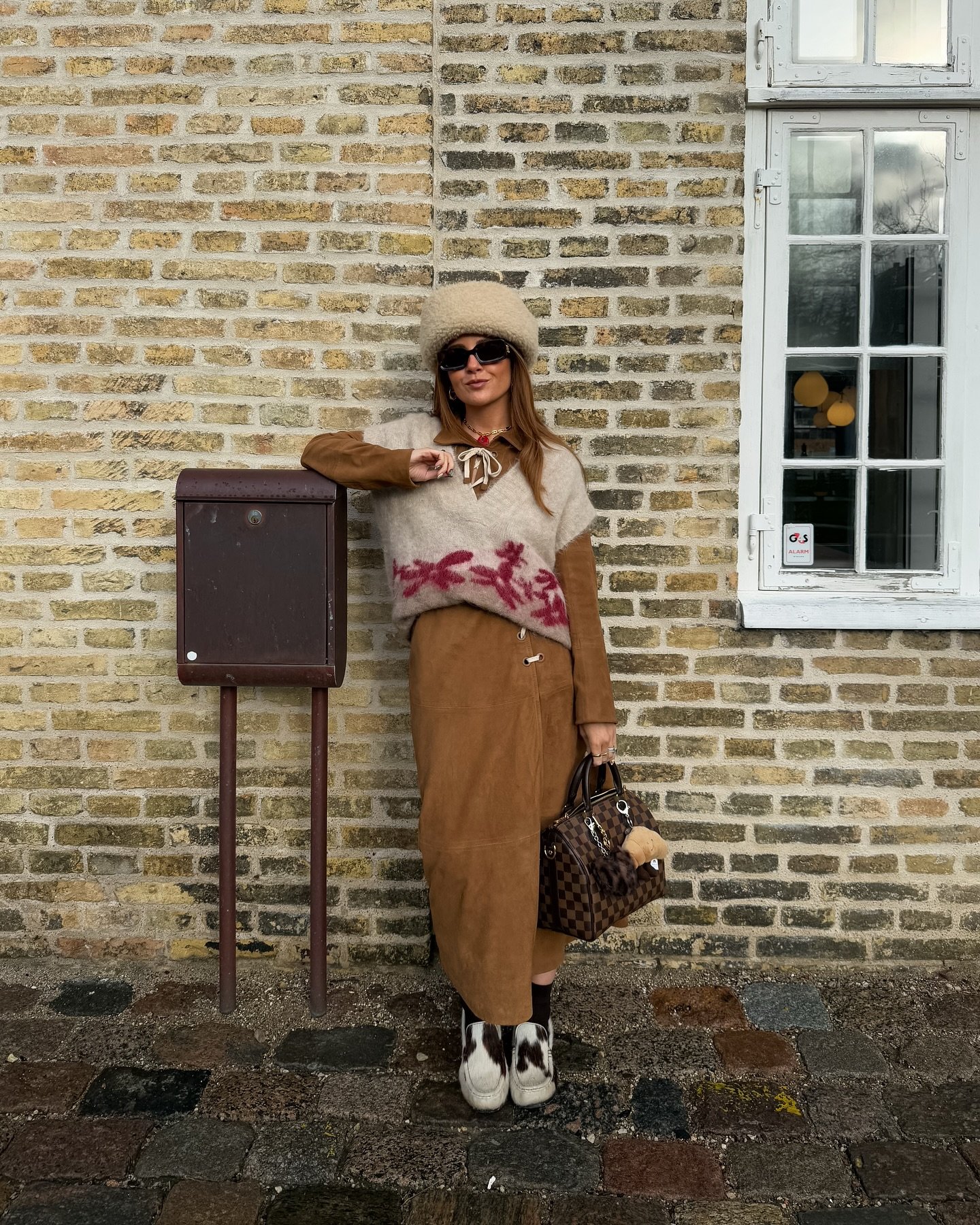 Mulher com chapéu e óculos escuros usa colete de lã e casaco marrom em estilo casual de inverno.