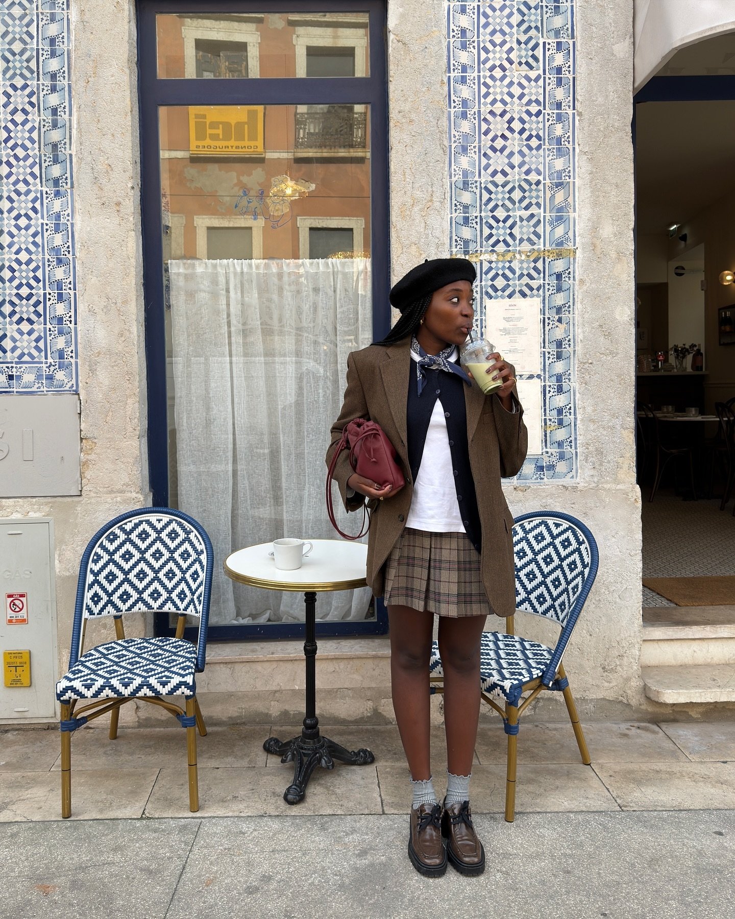 Pessoa com boina e saia xadrez, usando blazer, toma bebida em café ao ar livre. Looks de inverno com saia.