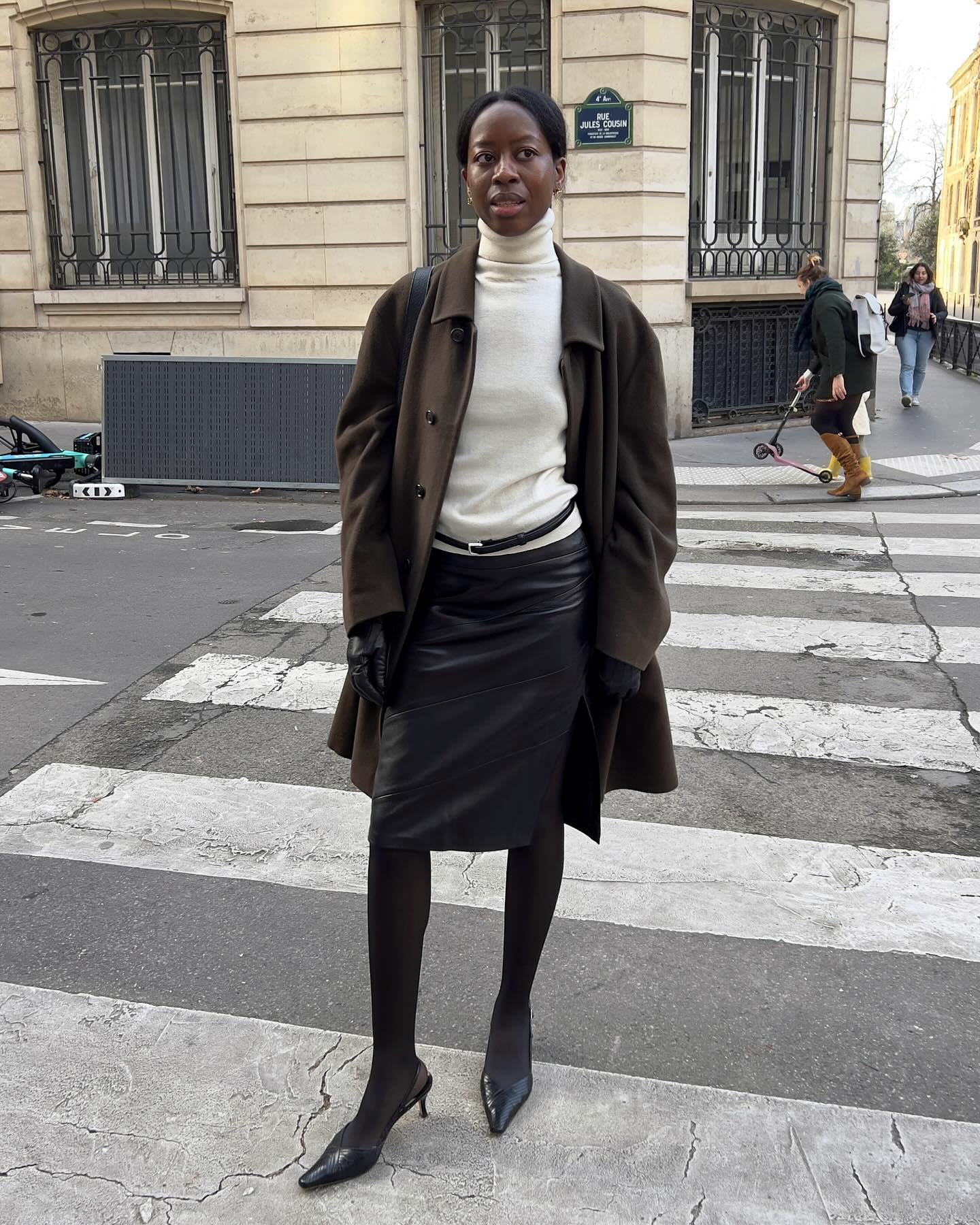 Pessoa com casaco marrom, saia preta e suéter branco cruza a rua, compondo um look de inverno elegante.