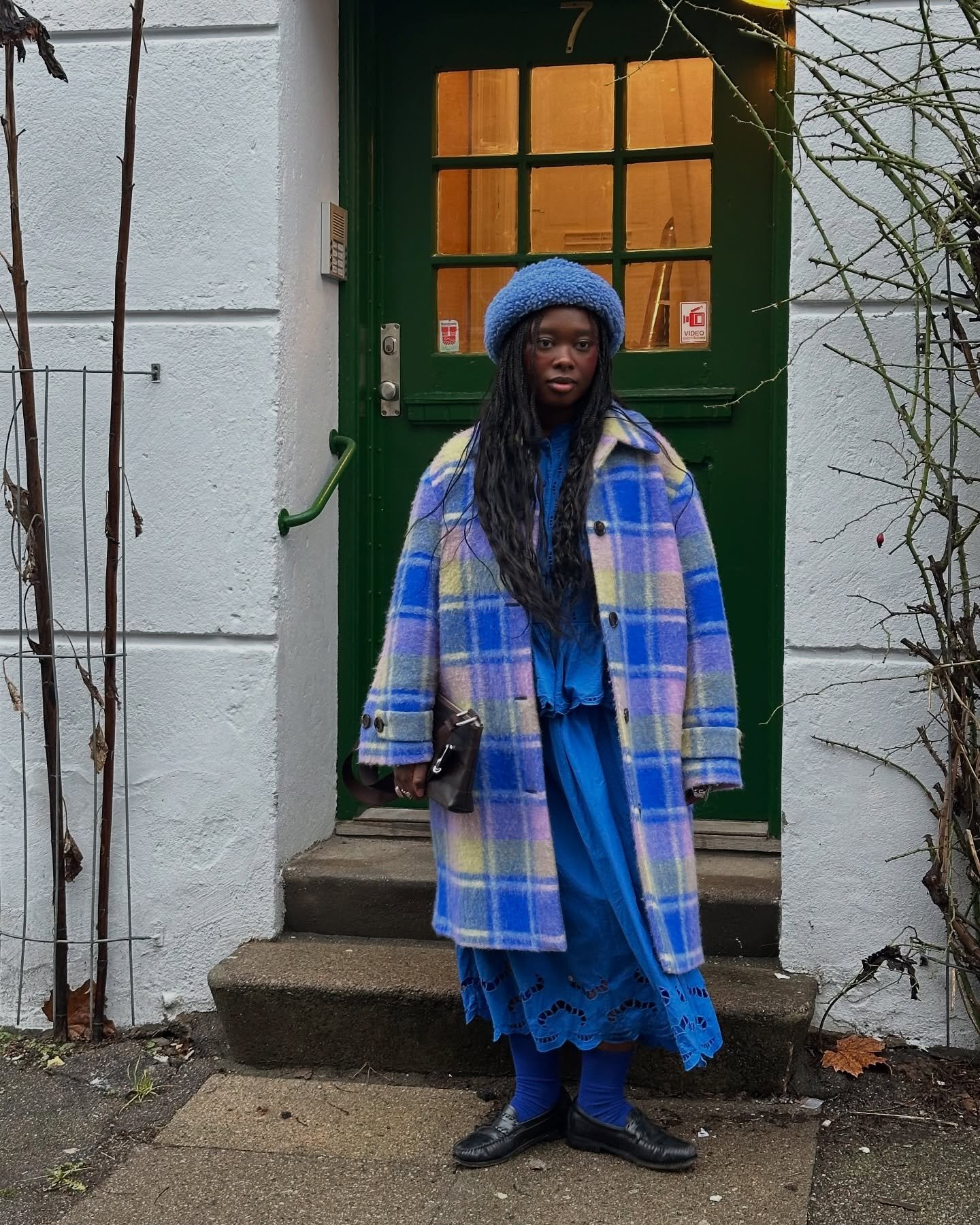 Casaco xadrez multicolorido sobre saia azul e chapéu em look de inverno frente a porta verde.