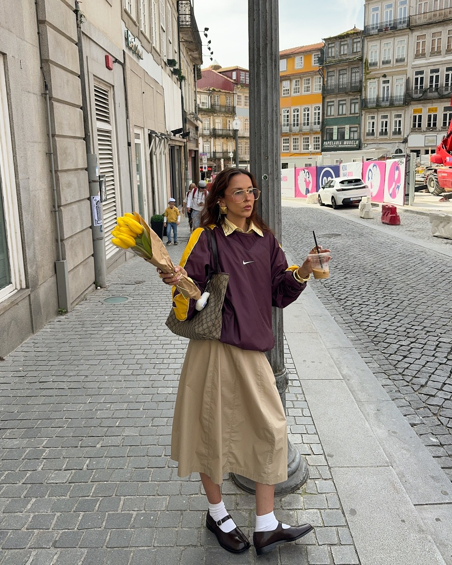 Pessoa caminhando em rua de paralelepípedos com saia longa bege, moletom roxo e segurando flores amarelas.
