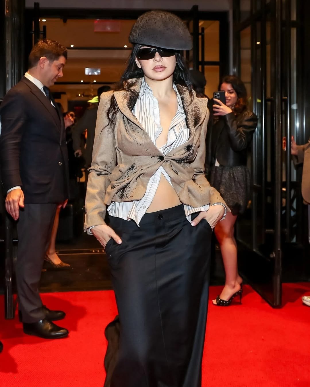 Pessoa em evento, de óculos escuros, chapéu e roupas elegantes em tapete vermelho, estilo after party de gala.