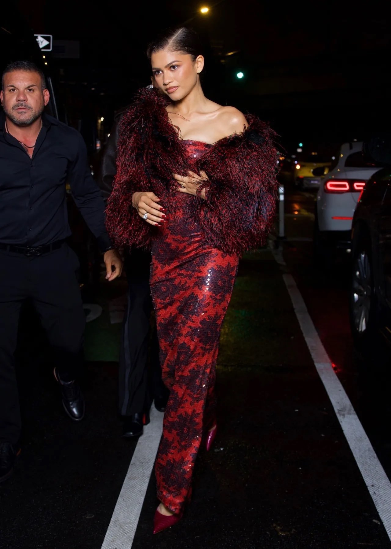 Vestido vermelho brilhante com detalhes de renda, combinado com um casaco de penas escuras, usado em festa pós-Met Gala 2025.