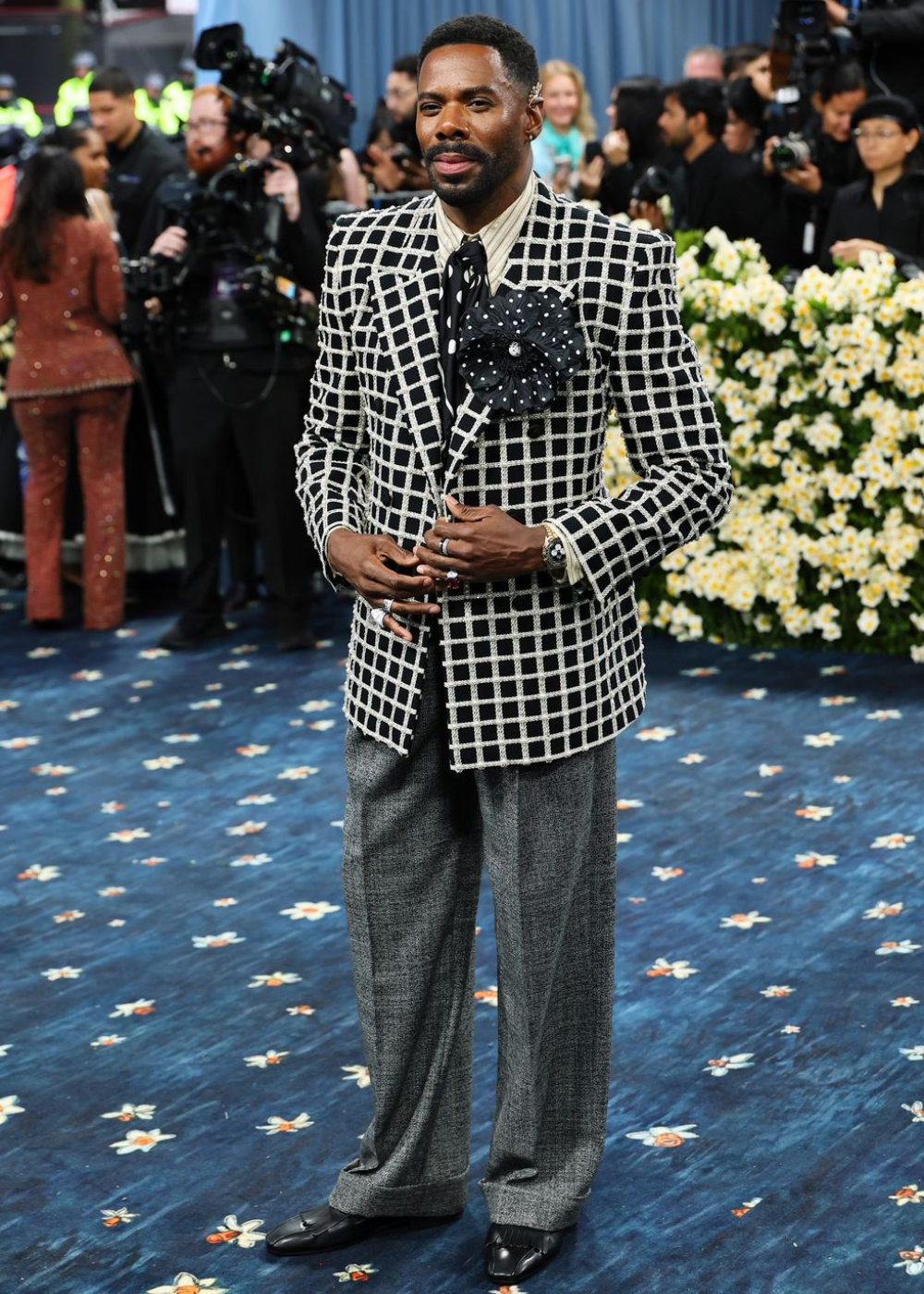 A pessoa está em um tapete azul com flores, usando blazer xadrez preto e branco, camisa listrada, gravata de bolinhas e flor decorativa.