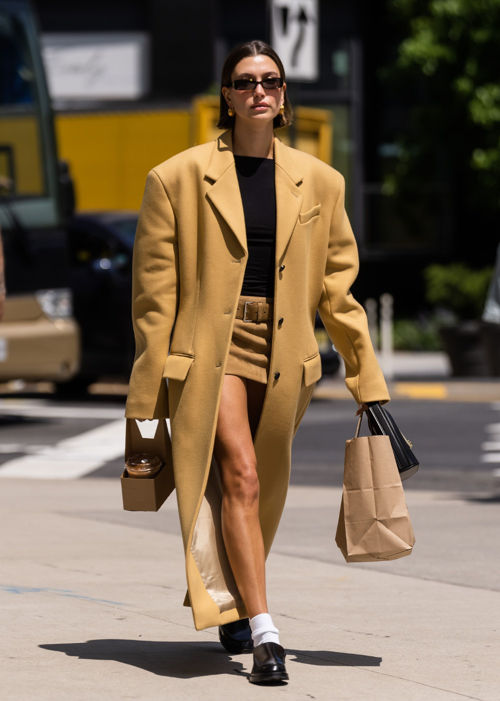Pessoa usando casaco amarelo, shorts, mocassim com meia branca, óculos escuros, carregando sacolas.