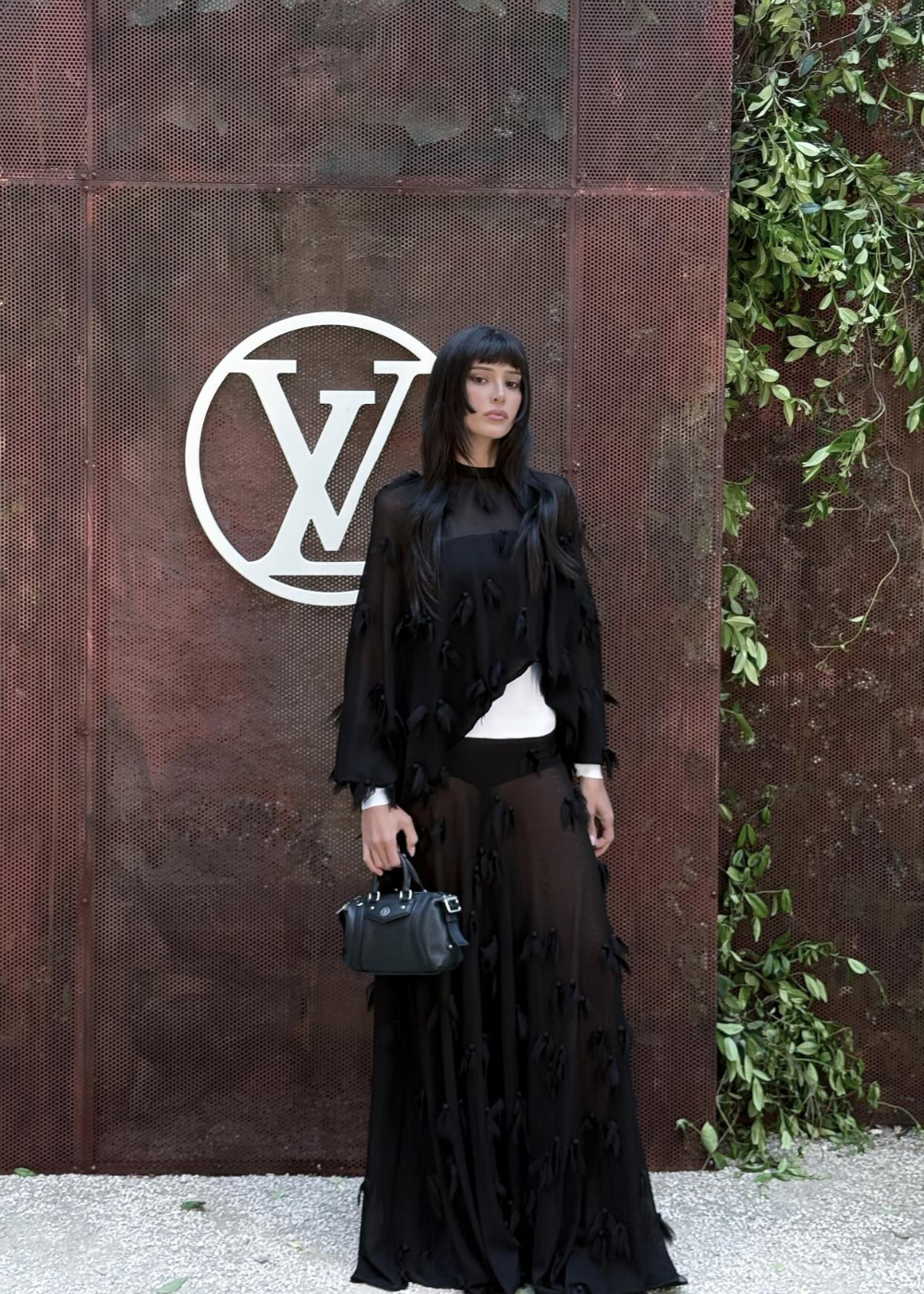 Modelo em frente ao logo LV, usando vestido preto com detalhes. Evento Cruise 2026.