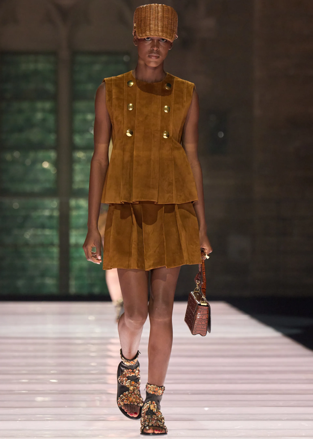 Desfile de moda, modelo com vestido ocre e bolsa marrom, exibindo coleção Cruise 2026.