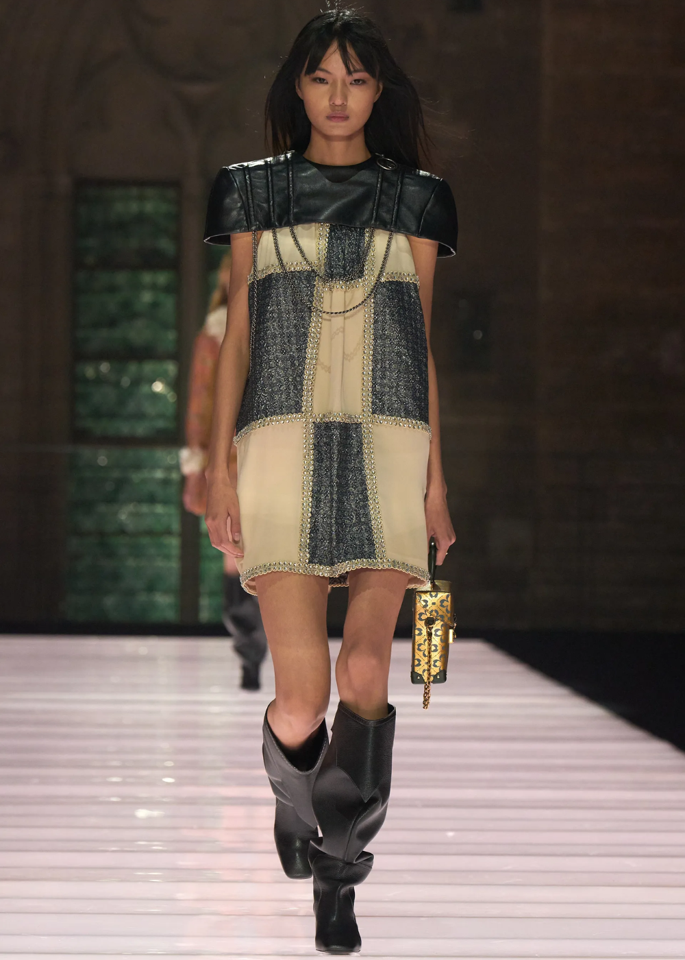 Modelo em desfile usando vestido com detalhes metálicos, capa de couro e botas altas. Ela carrega uma bolsa dourada.