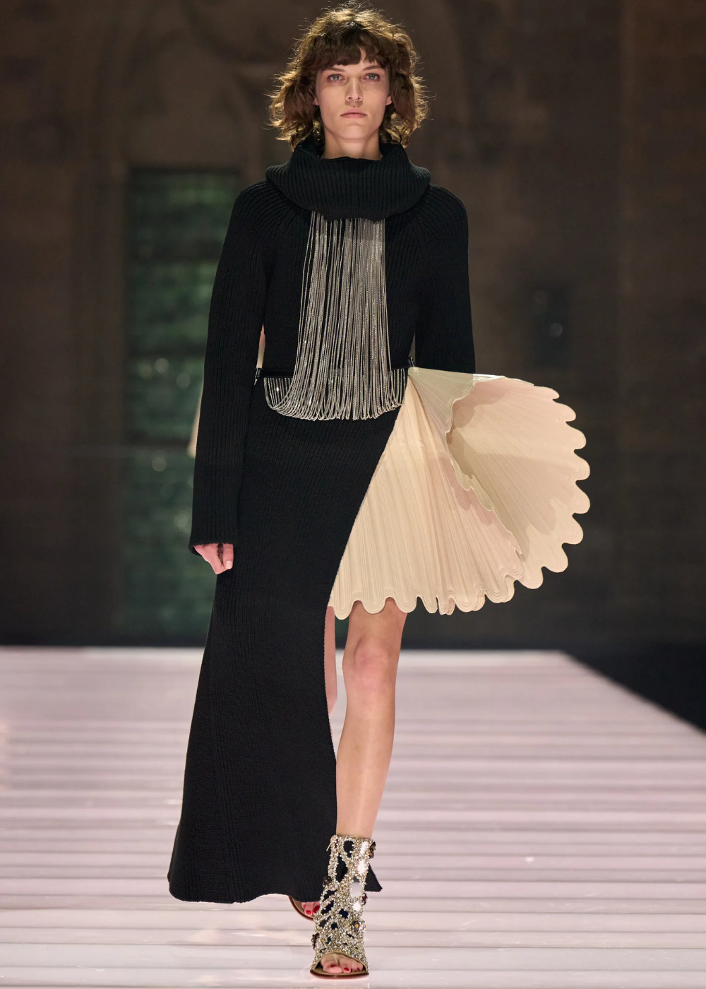 Modelo em desfile com vestido assimétrico preto e bege, destaque para franjas metálicas. Destaque do evento Cruise 2026.