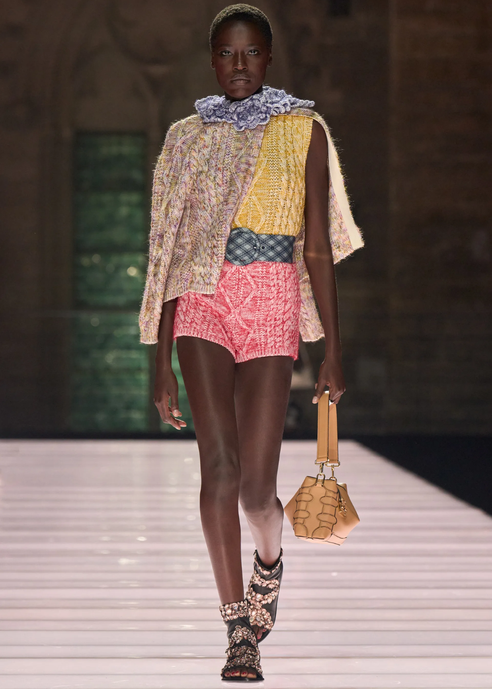 Modelo vestindo blusa texturizada amarelo e lilás, shorts rosa, cinto azul e bolsa bege, desfilando na coleção Cruise 2026.