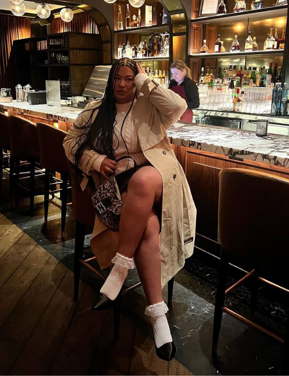 Mulher sentada em um bar elegante, usando casaco bege e meia de babado, enquanto toca o cabelo.