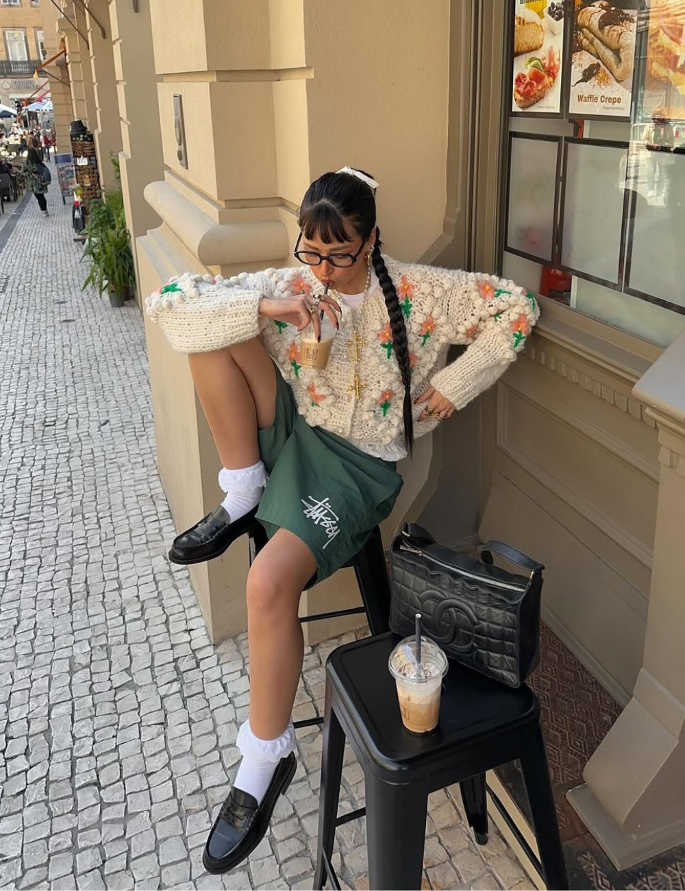 Pessoa sentada em banco, usando suéter e meia de babado, bebendo café gelado. Mochila ao lado sobre o banco.
