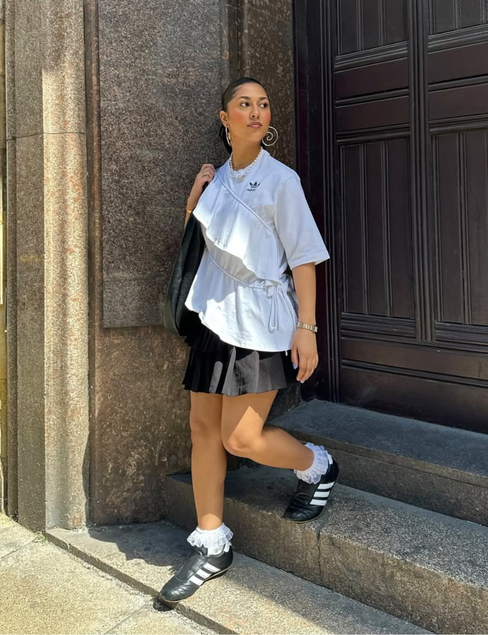 Mulher com camiseta branca e saia preta, usando meia de babado e tênis, encostada em uma parede de pedra.