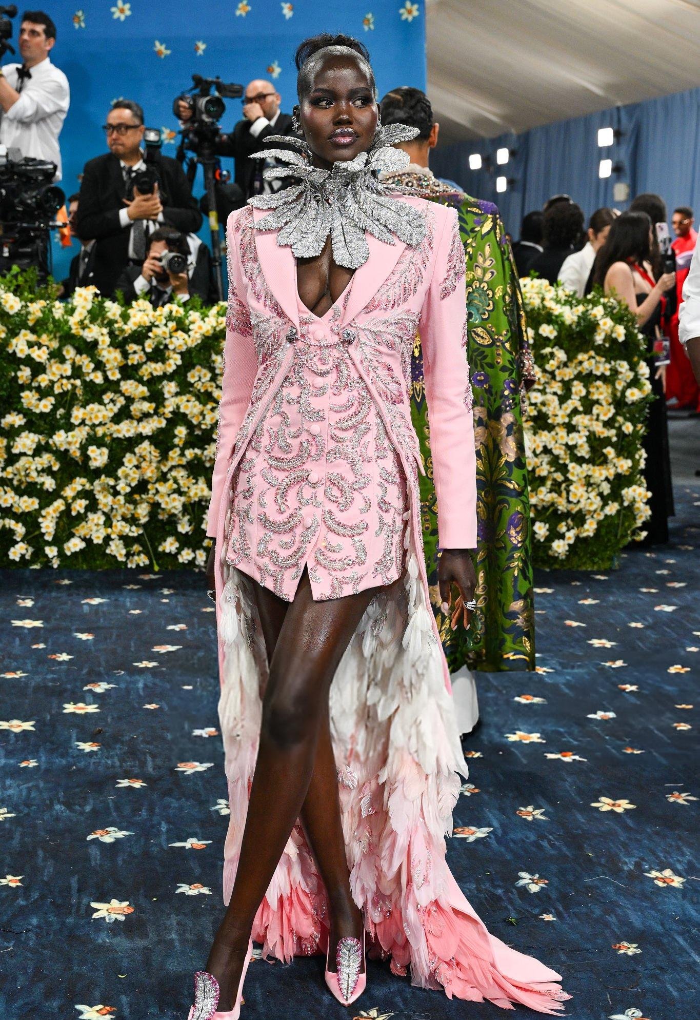 Desfile de moda no Met Gala 2025, com vestido rosa detalhado, gola extravagante e fundo floral.