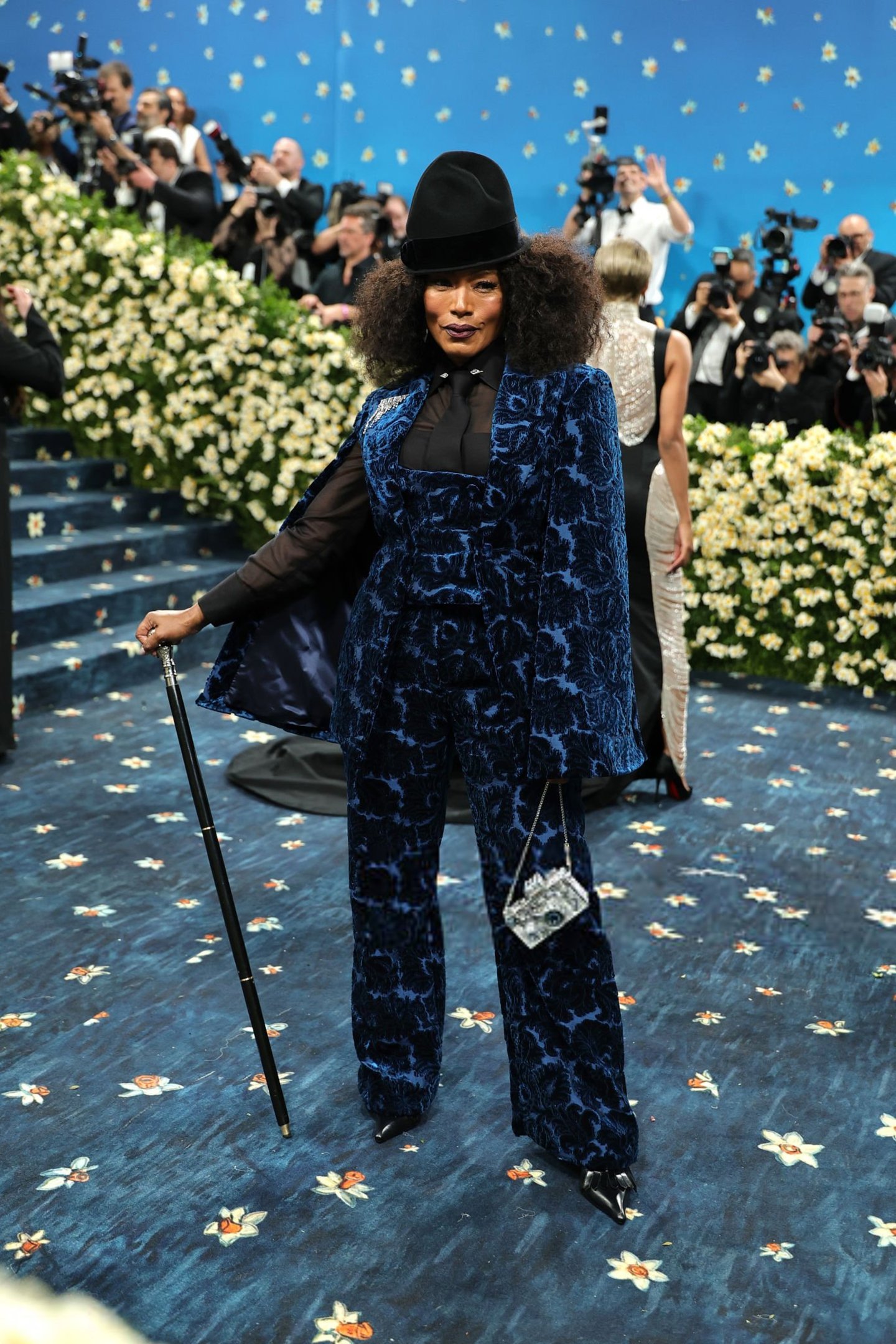 Celebridade com terno azul floreado e bengala no Met Gala 2025. Tapete azul com flores.