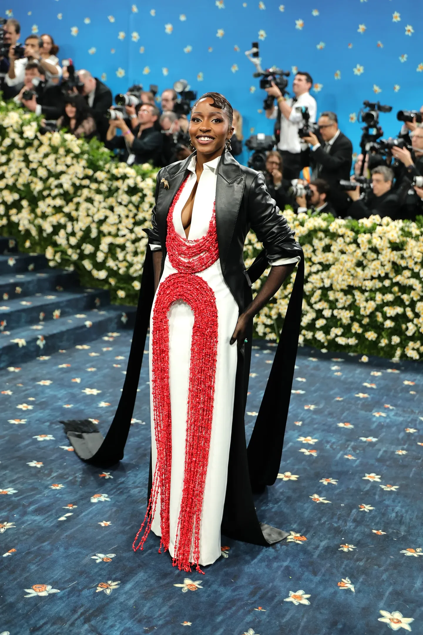 Participante exibe vestido vermelho e branco com capa preta em evento elegante, cercada por fotógrafos. Met Gala 2025.