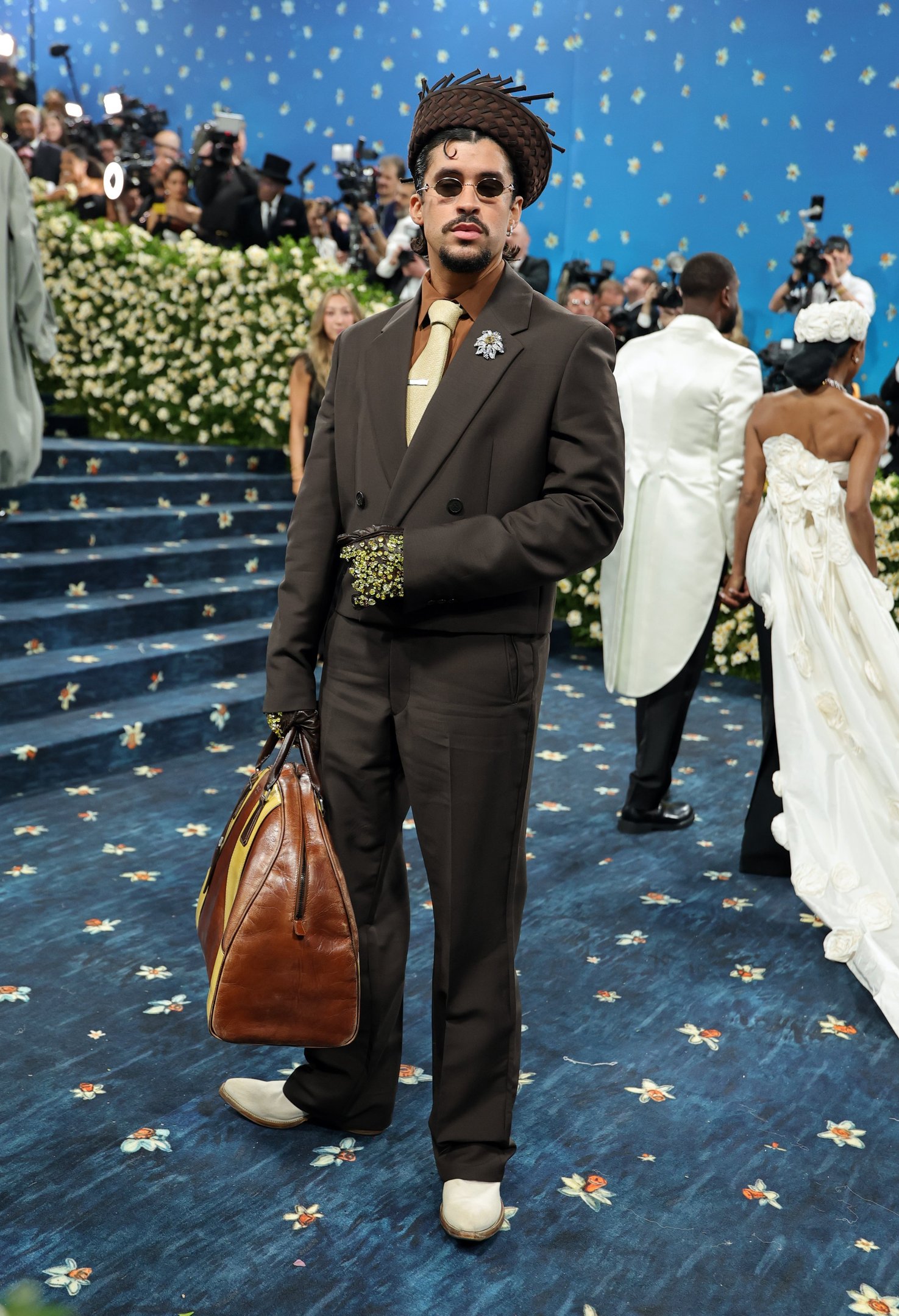 Homem em terno marrom com acessórios elegantes no Met Gala 2025, segurando bolsa grande. Fundo de escadaria florida.