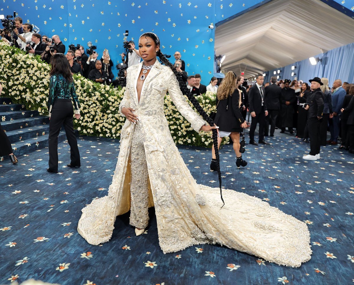 Celebridade com vestido ornamentado em evento glamoroso, Met Gala 2025, rodeada por fotógrafos e decoração floral.