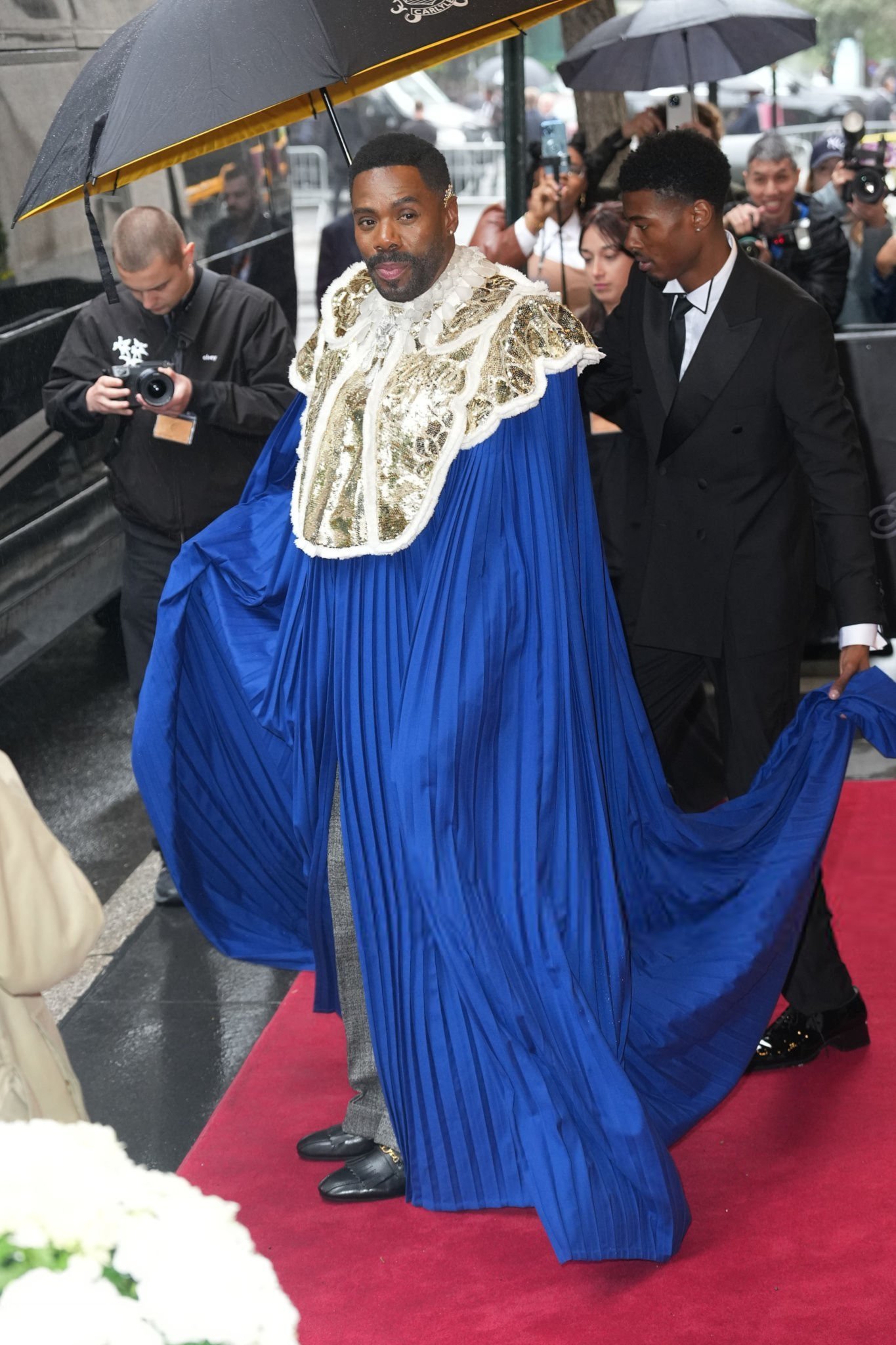 Pessoa em capa azul e dourada sob guarda-chuva, no tapete vermelho do Met Gala 2025.