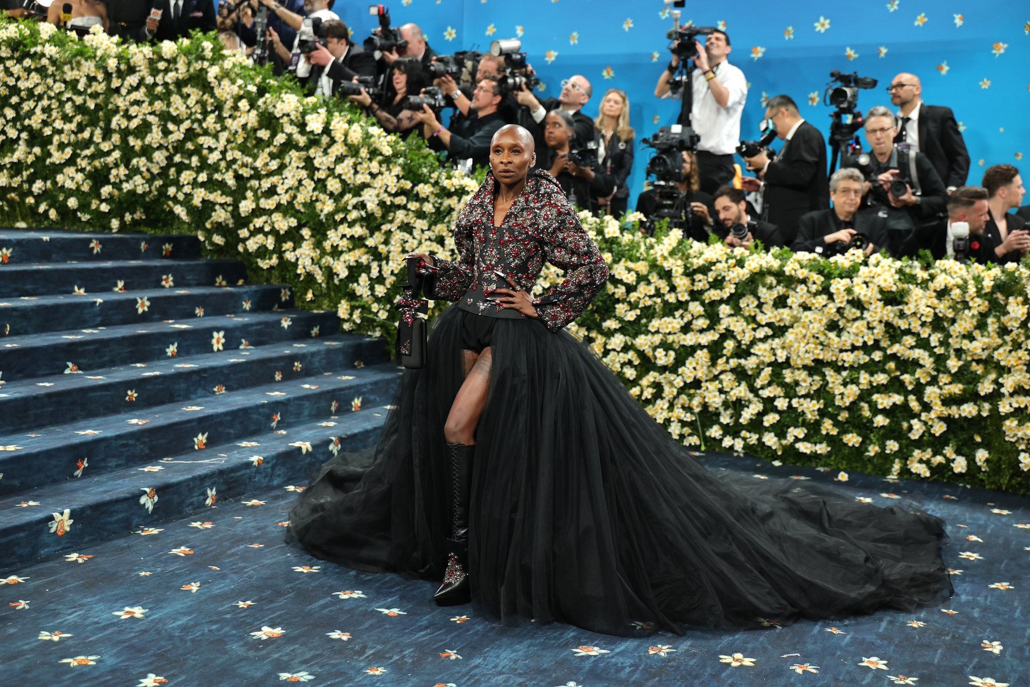 Celebridade usando vestido preto elegante com cauda longa no Met Gala 2025, cercada por fotógrafos e escadaria florida.
