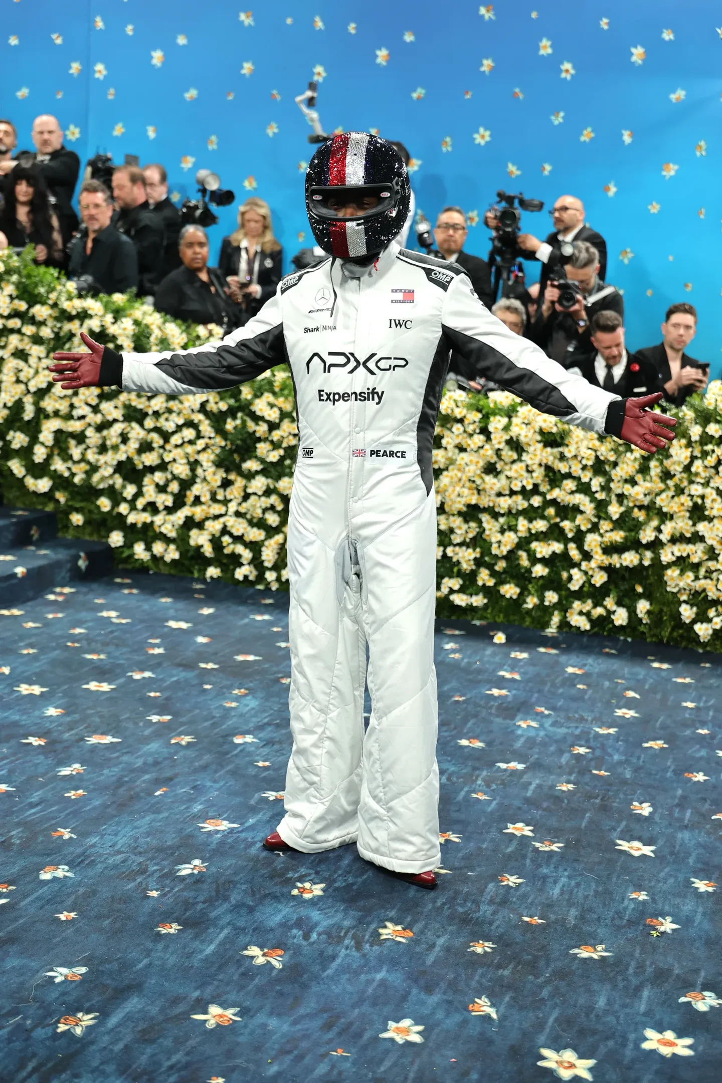 Participante no Met Gala 2025 veste macacão e capacete de automobilismo decorado. Fundo com flores e público.