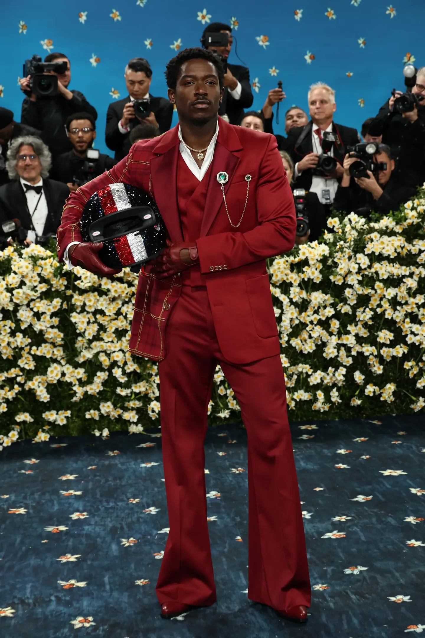 Homem com terno vermelho, camisa branca e broche, segura capacete estilizado. Ao fundo, fotógrafos e flores. Looks masculinos