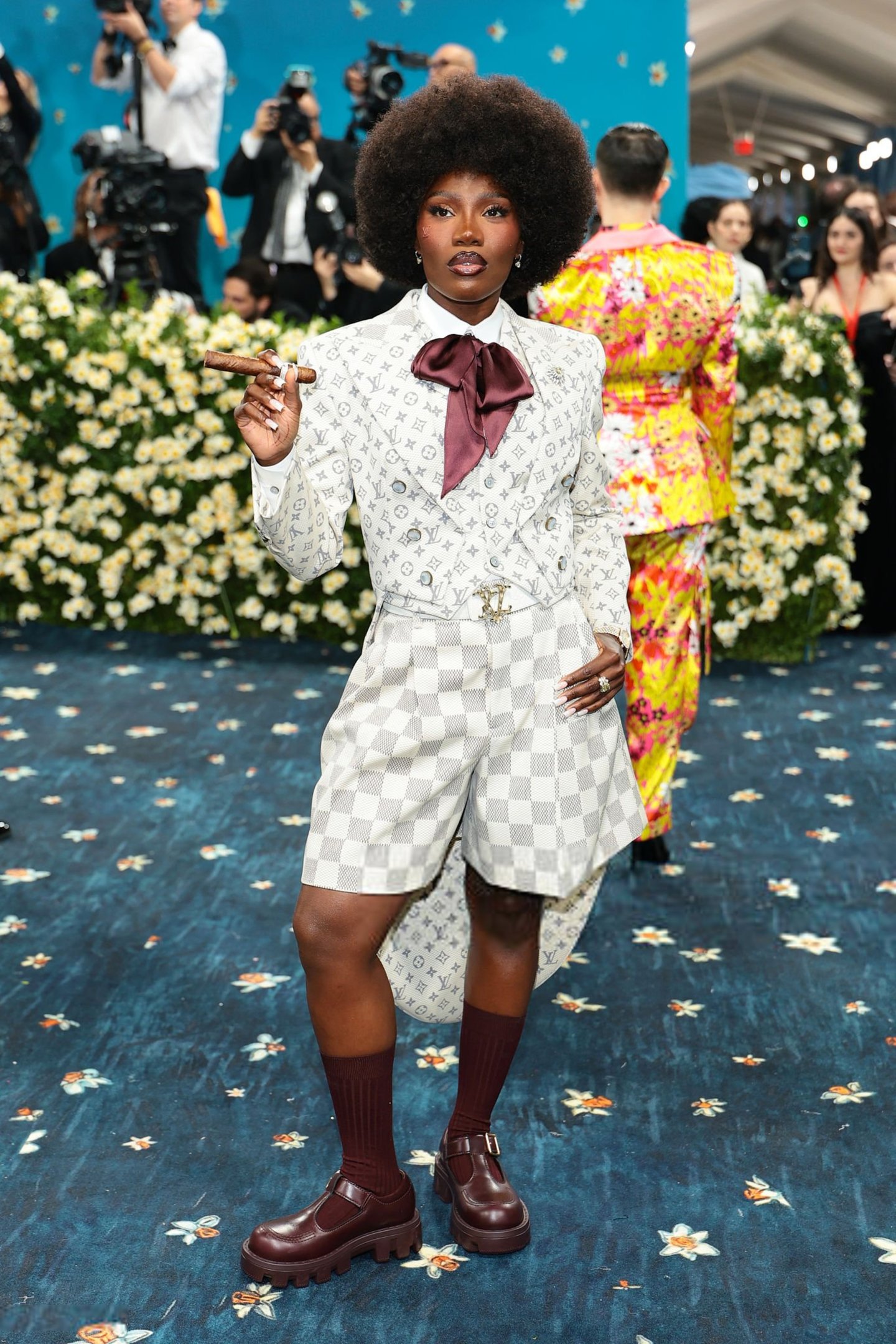 Pessoa vestida de alfaiataria estampada, com gravata bordô, em evento de moda Met Gala 2025. Ela segura um charuto.