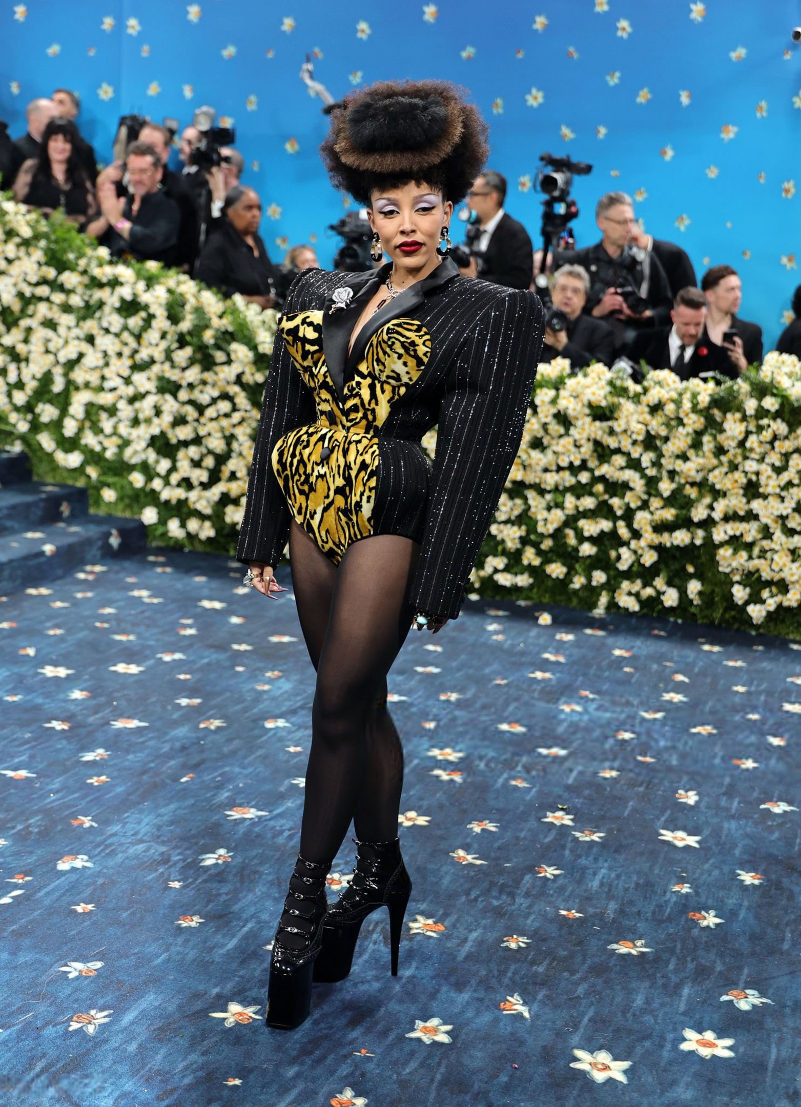 Celebridade em traje preto e dourado ousado com padronagem tigrada, em evento de tapete azul com flores.