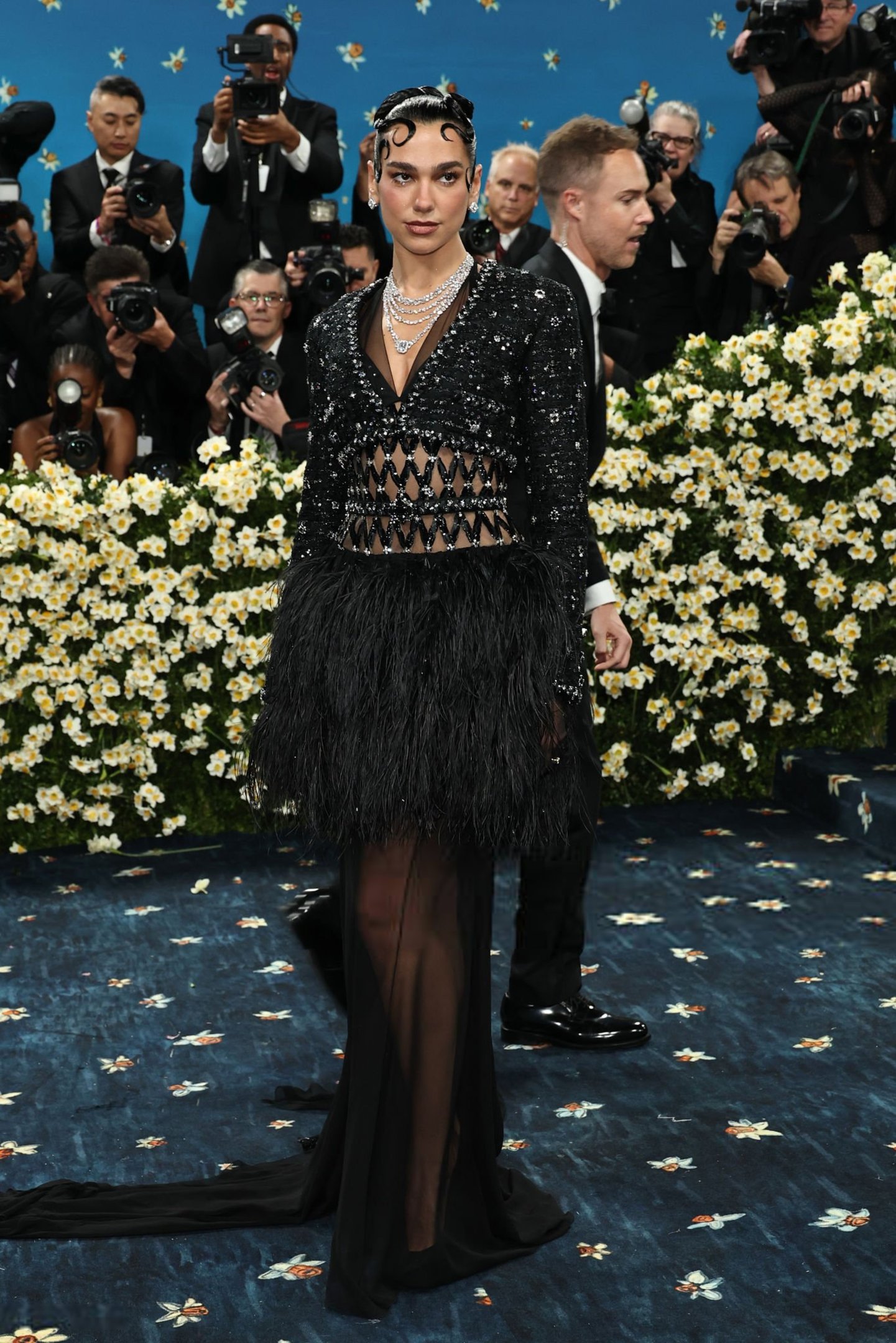Celebridade em evento com vestido preto elegante e aplique de penas, cercada por fotógrafos no Met Gala 2025.