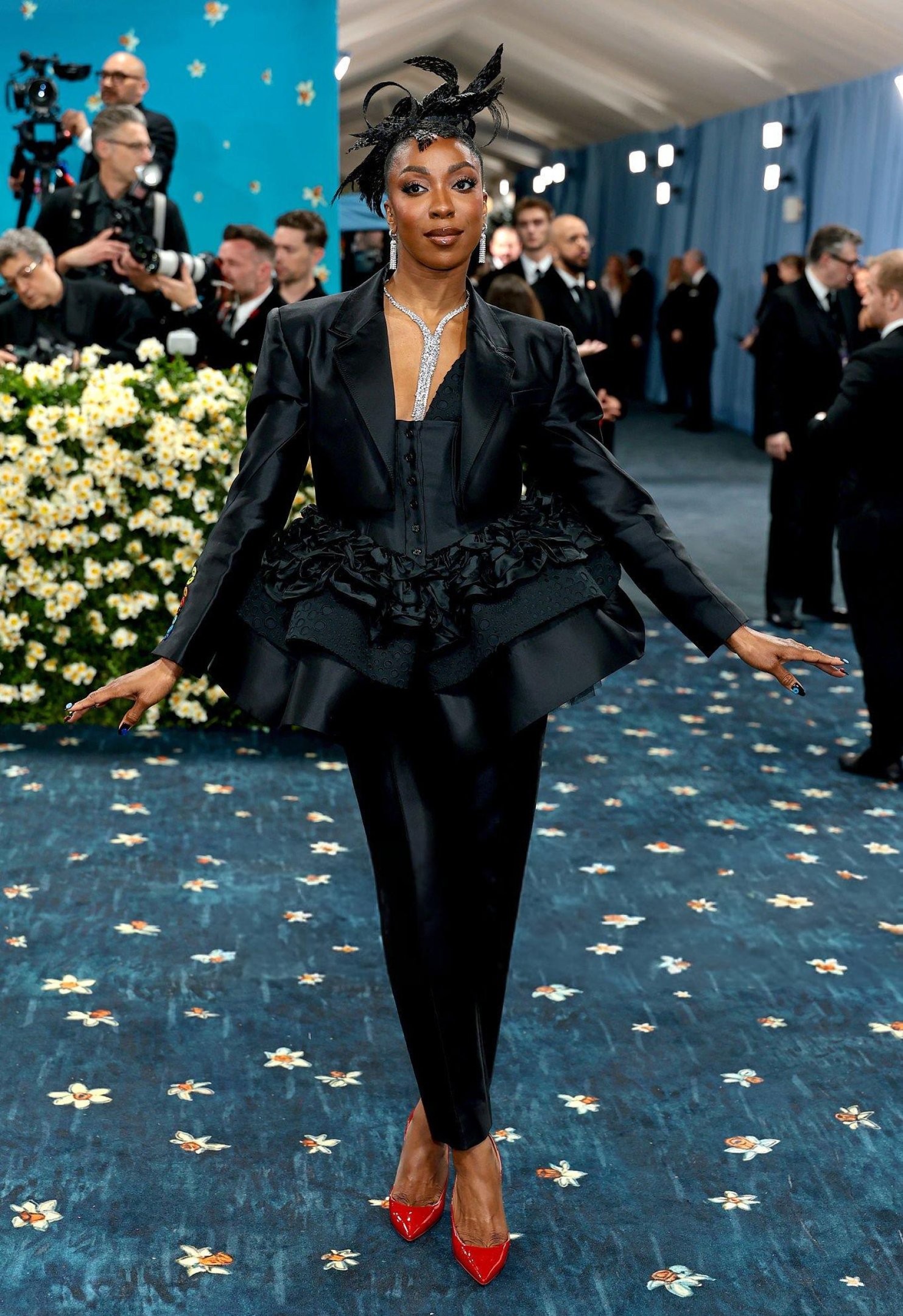 Pessoa elegante em traje preto adornado por plumas e sapatos vermelhos no Met Gala 2025.