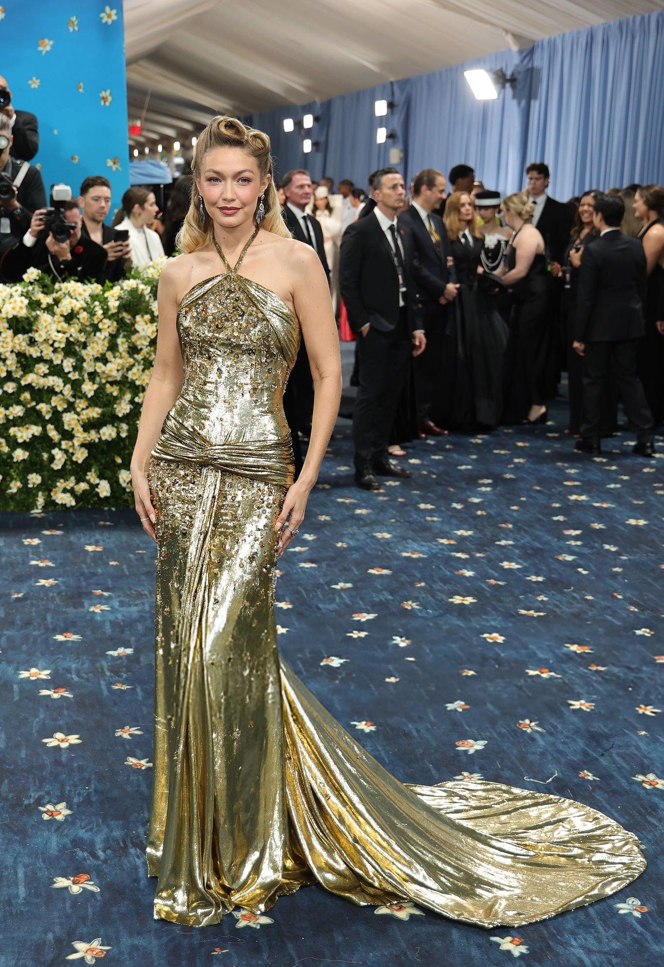 Pessoa usando vestido dourado brilhante no Met Gala 2025, rodeada por convidados elegantes.