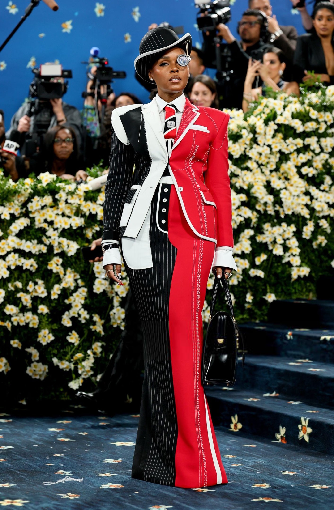 Pessoa em traje formal preto, branco e vermelho, com risca de giz. Fundo de flores e fotógrafos ao redor.