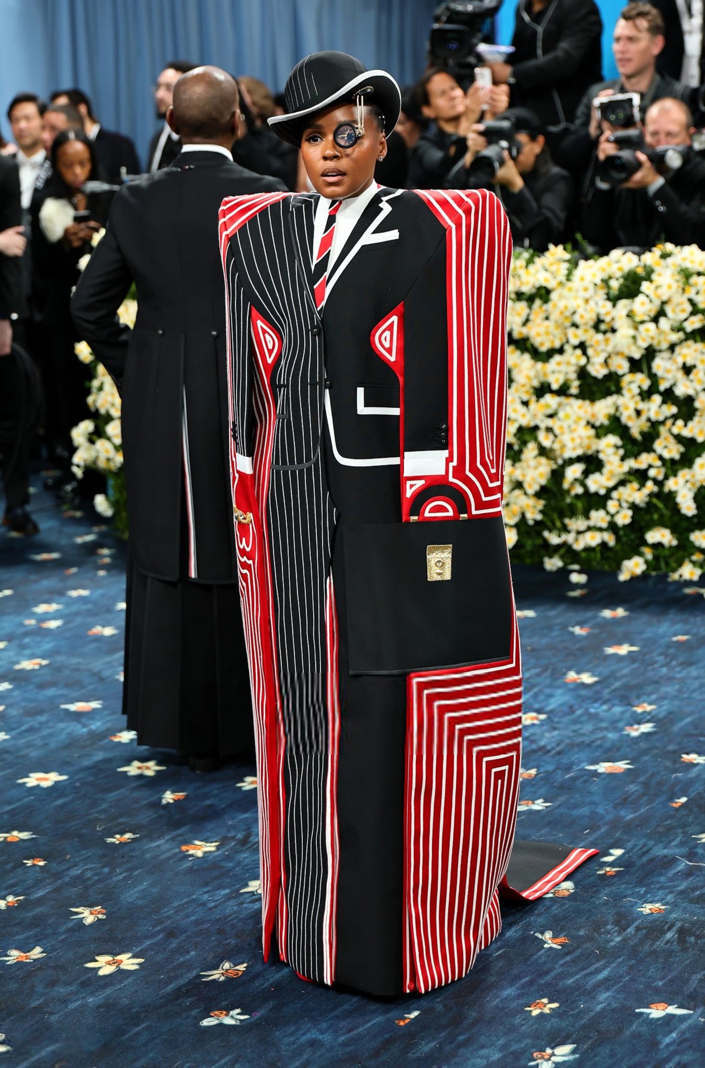 Pessoa com chapéu e monocel em traje geométrico preto, vermelho e branco no Met Gala 2025, posando em tapete azul.