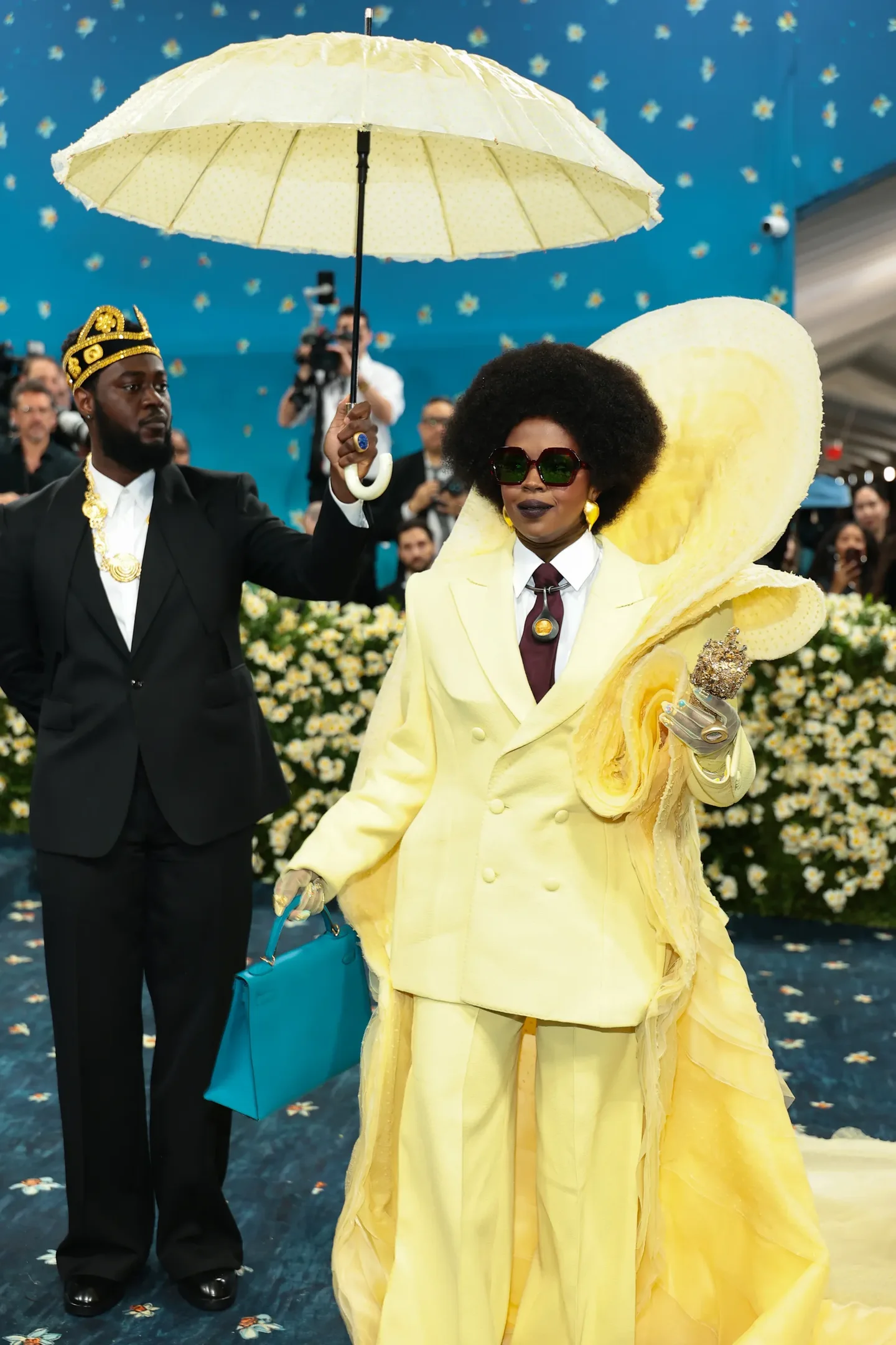 Figuras em roupas amarelas e pretas elegantes com guarda-chuva e bolsa azul no Met Gala 2025. Fundo floral.
