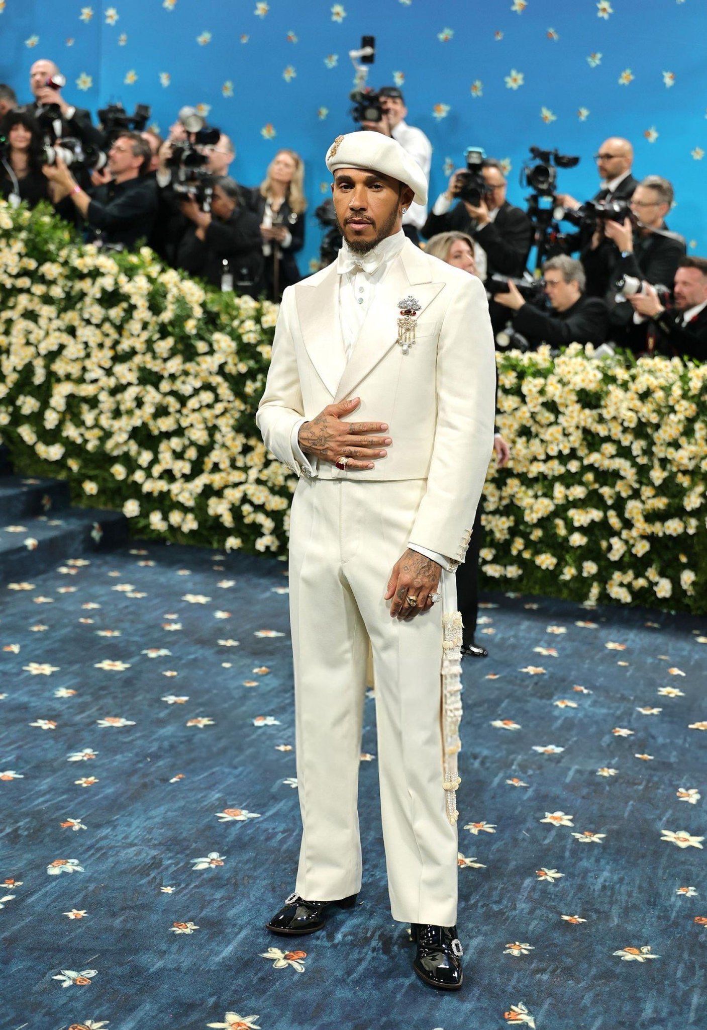 Elegante traje branco com boina e acessórios, em evento estilo Met Gala 2025, piso e fundo floridos.