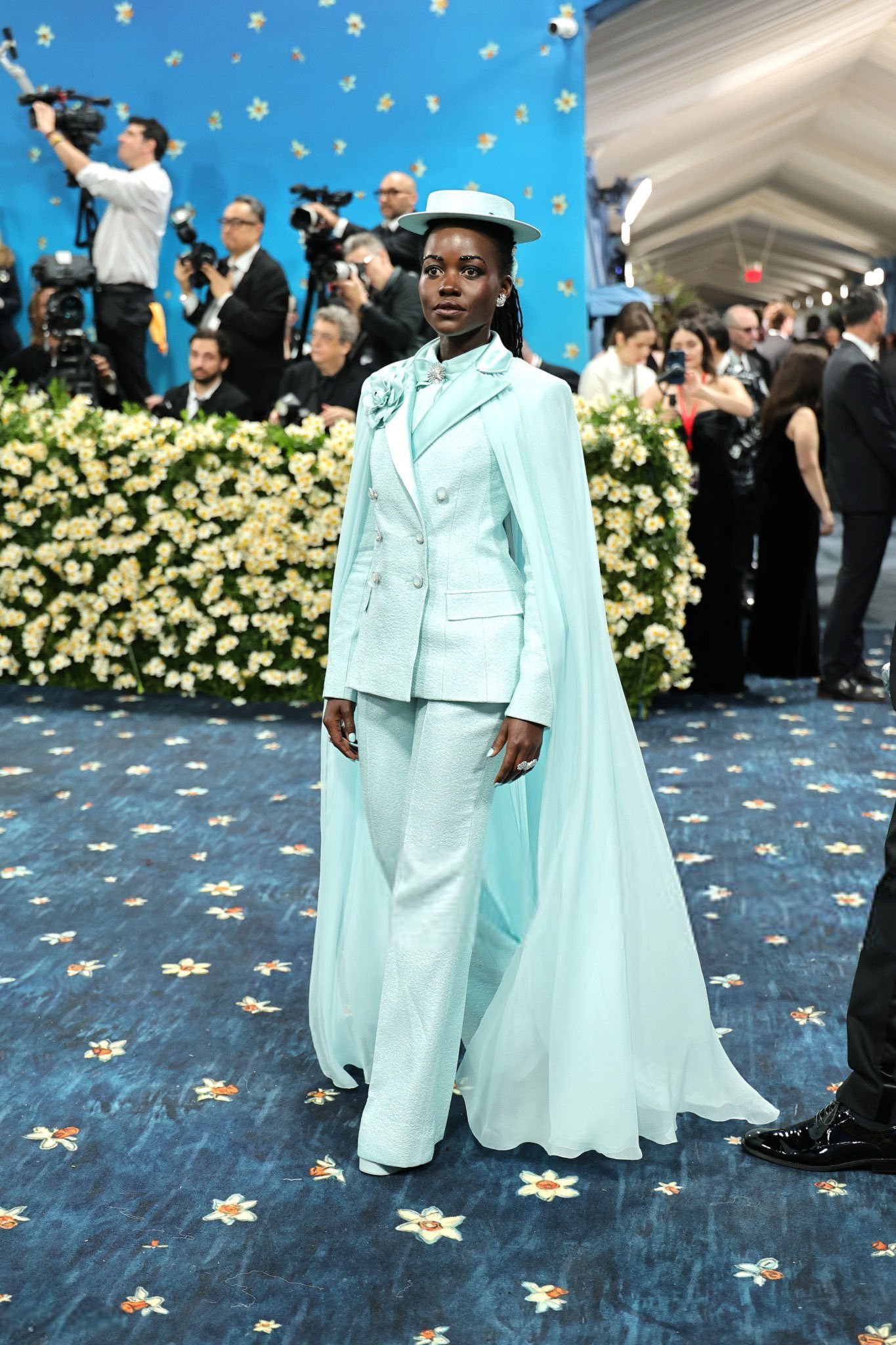 Pessoa em traje azul claro com capa longa no Met Gala 2025, rodeada de fotógrafos e flores amarelas.