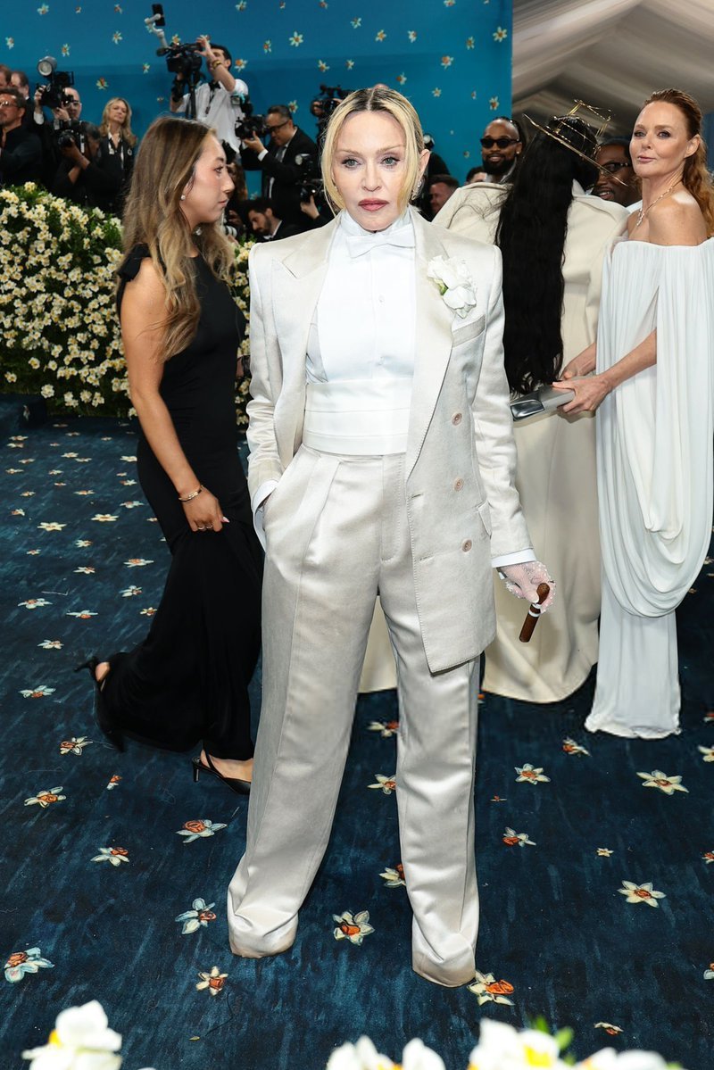 Celebridade em terno branco no tapete azul do Met Gala 2025, cercada por fotógrafos e convidados elegantes.