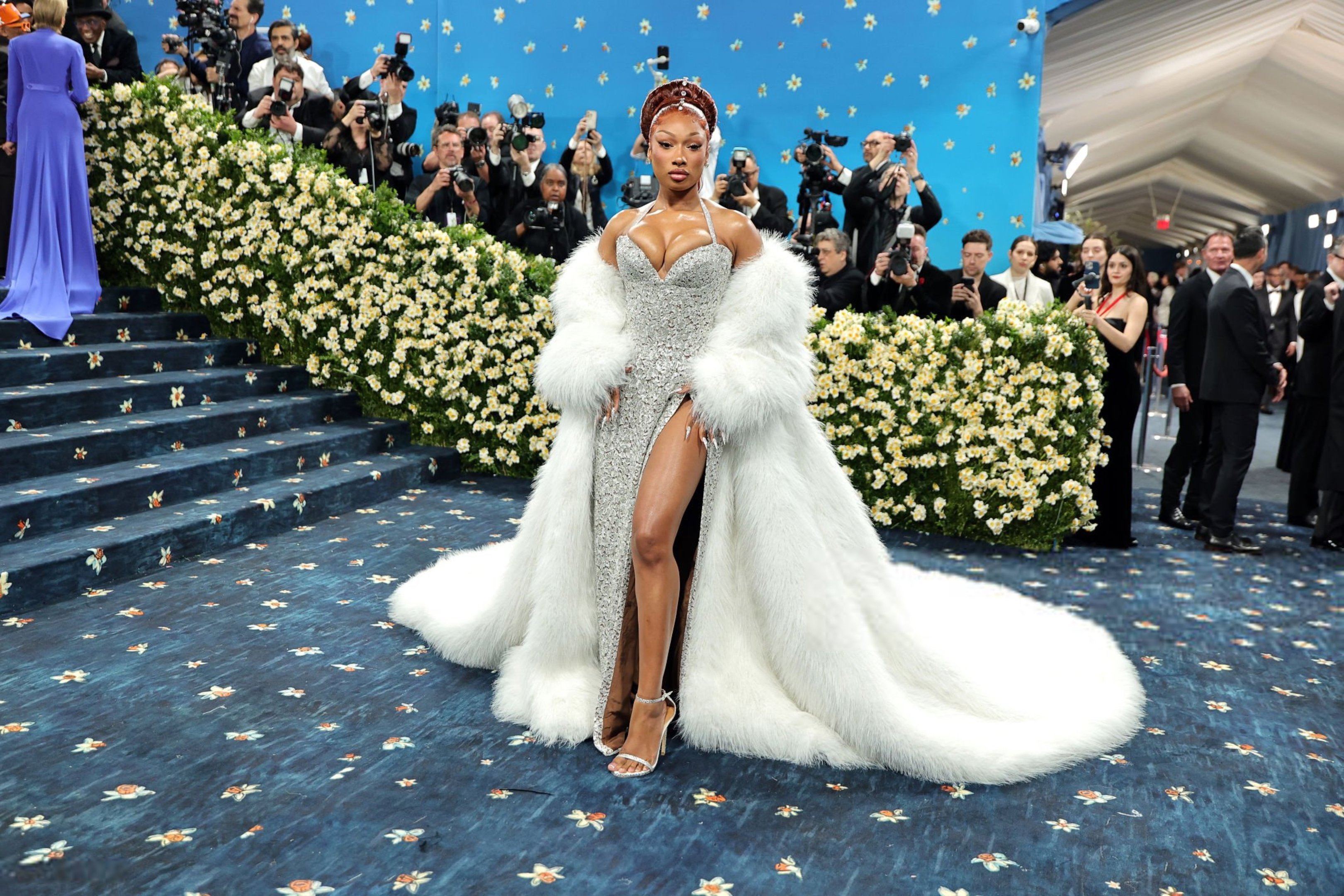 Celebridade em vestido prateado e casaco branco glamouroso, cercada por fotógrafos no Met Gala 2025.